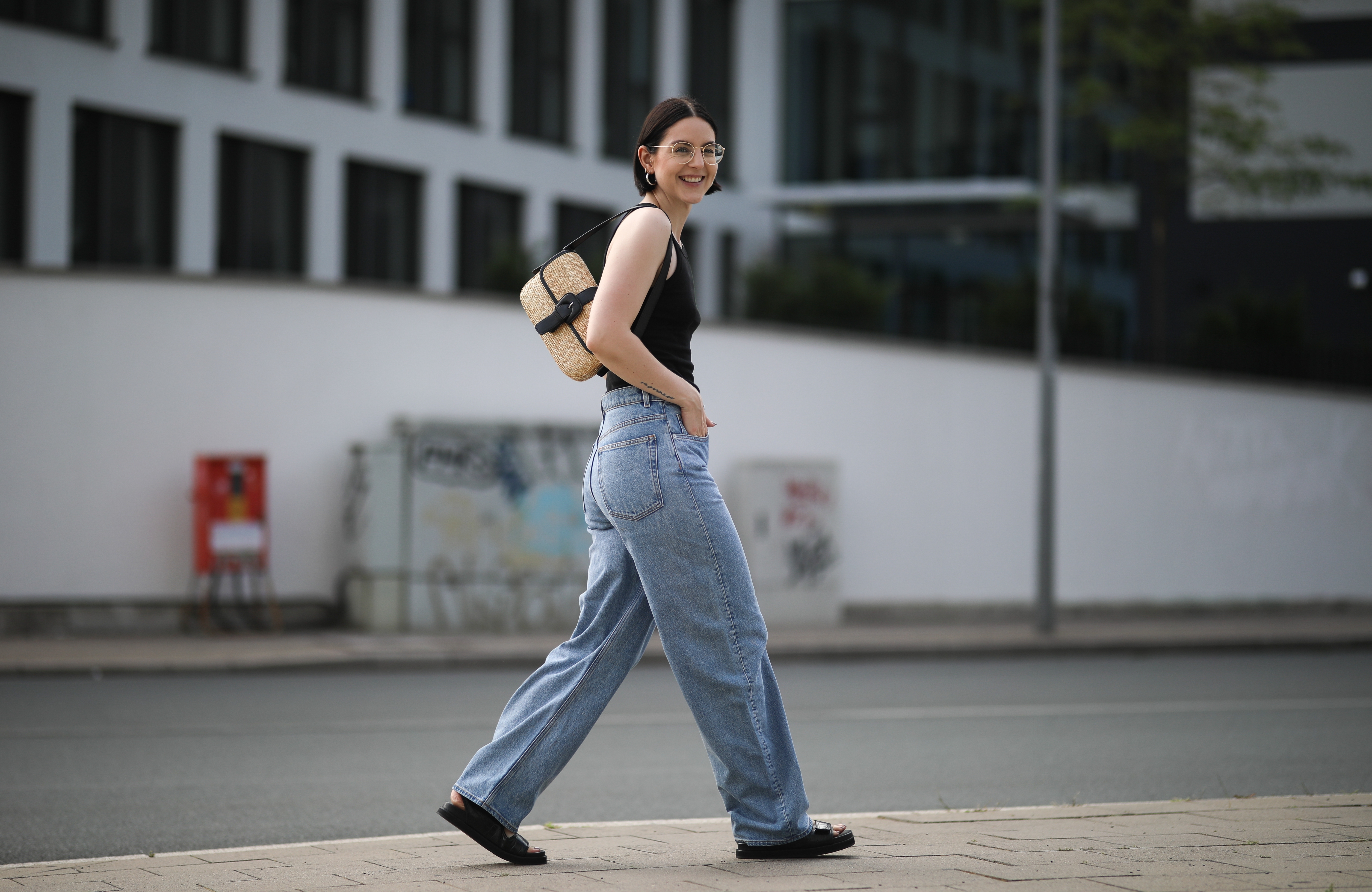 Person in casual outfit—sleeveless top, loose jeans, sandals—walks on a city street, carrying a small bag.