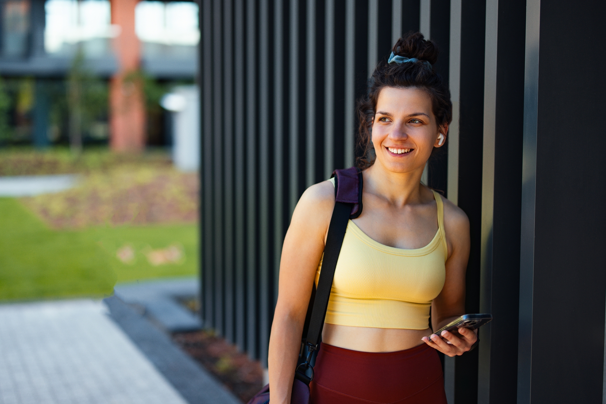 Woman in a workout outfit leans against a wall, smiling and holding a phone, with earbuds in. She's outdoors in a modern urban setting