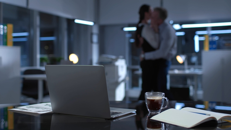 Two people shares an intimate moment in a dimly lit office, with a laptop, coffee, and notebook in the foreground