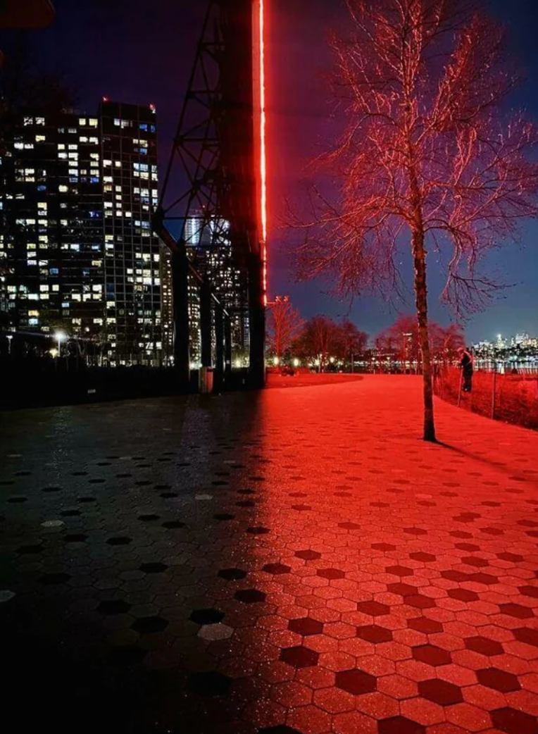 Night scene of a cityscape with a brightly lit red structure casting reflections on a patterned pathway, and illuminated buildings in the background