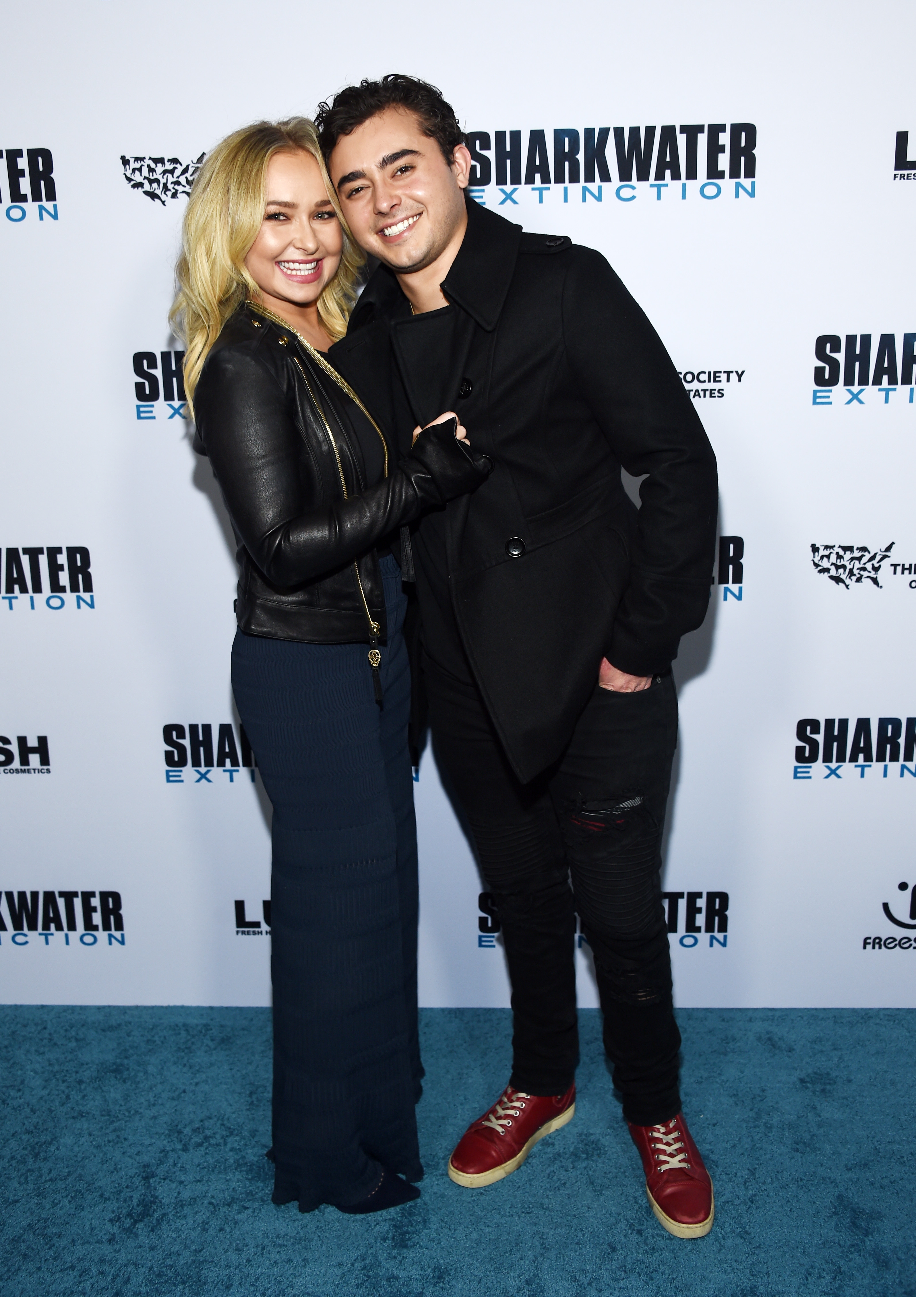 Two people smiling, posing together on a red carpet at the Sharkwater Extinction event; one wears a leather jacket, the other a dark coat