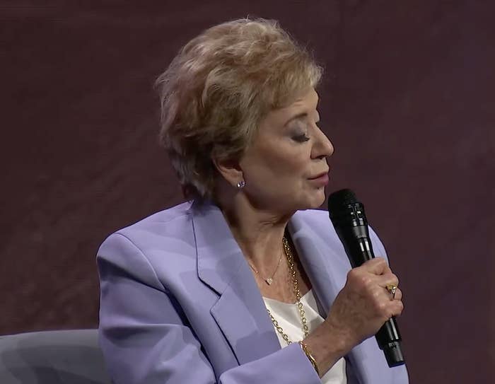 An older woman speaks into a microphone during a seated interview, wearing a professional blazer and gold necklace