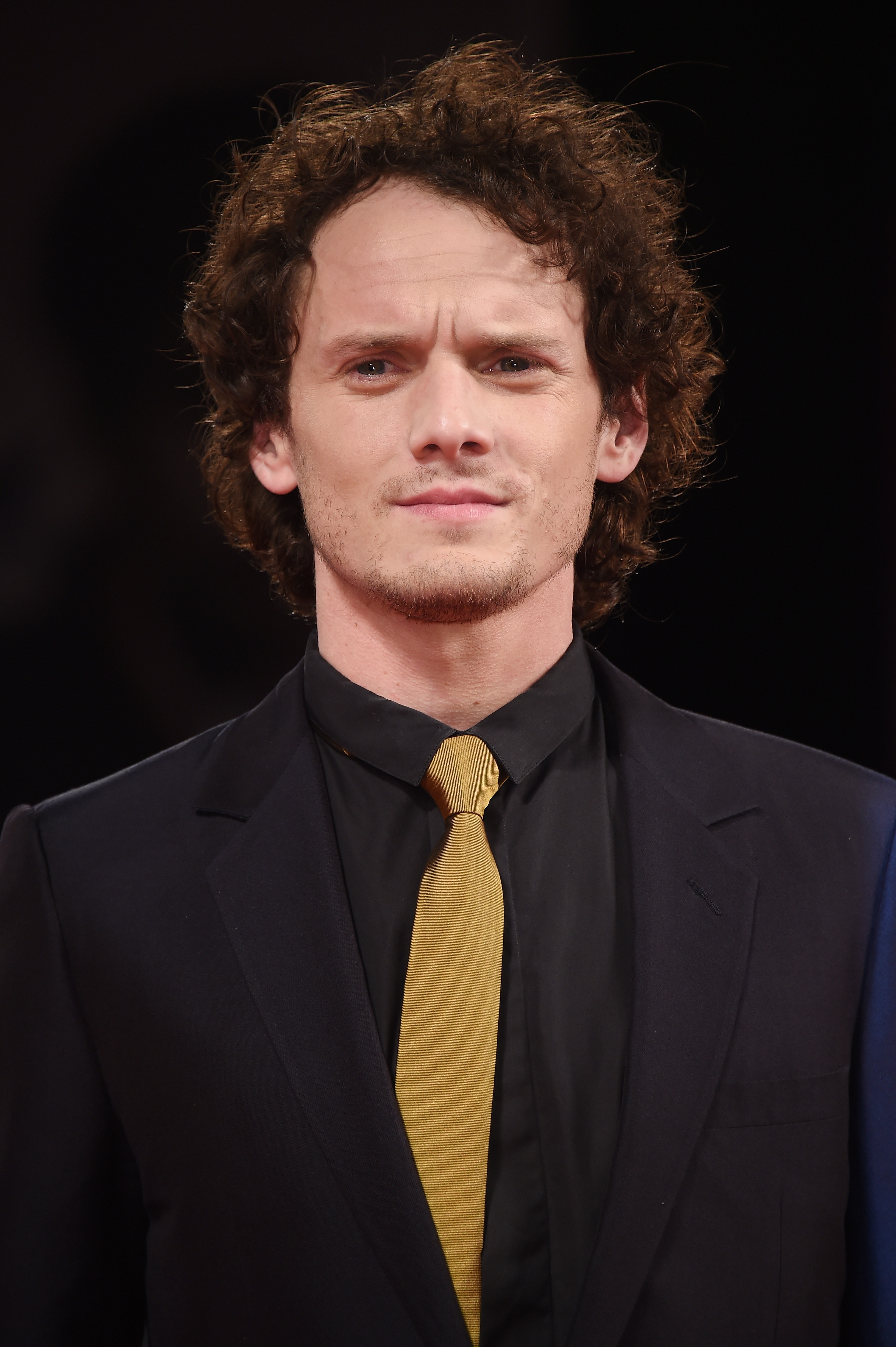 Person in a suit with a distinct hairstyle stands confidently, wearing a black shirt and mustard tie