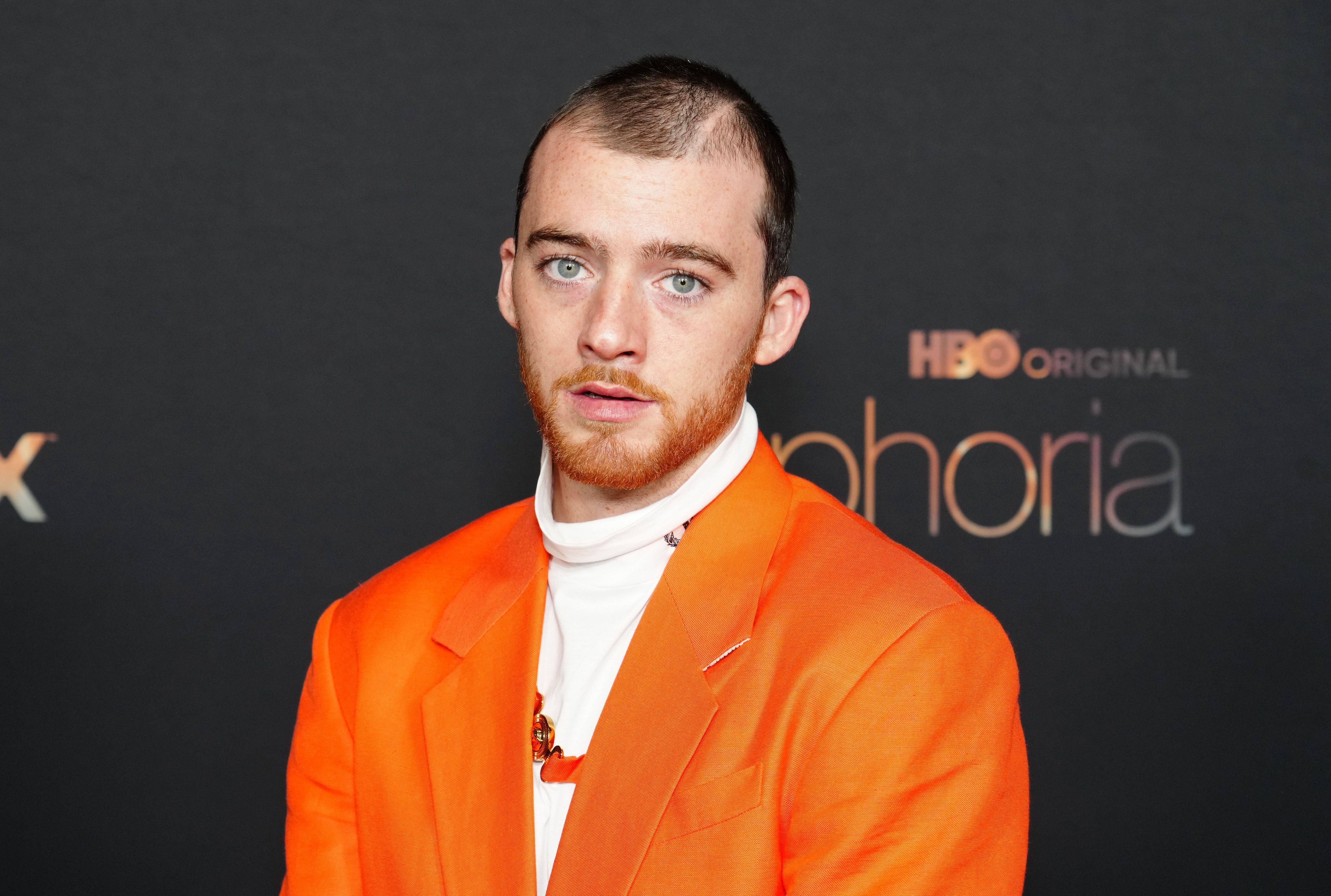 Person in formal orange jacket over turtleneck at an HBO event backdrop