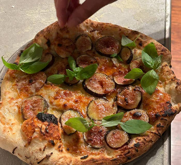 Hand reaching for a slice of pizza topped with figs, basil, and melted cheese on a baking tray