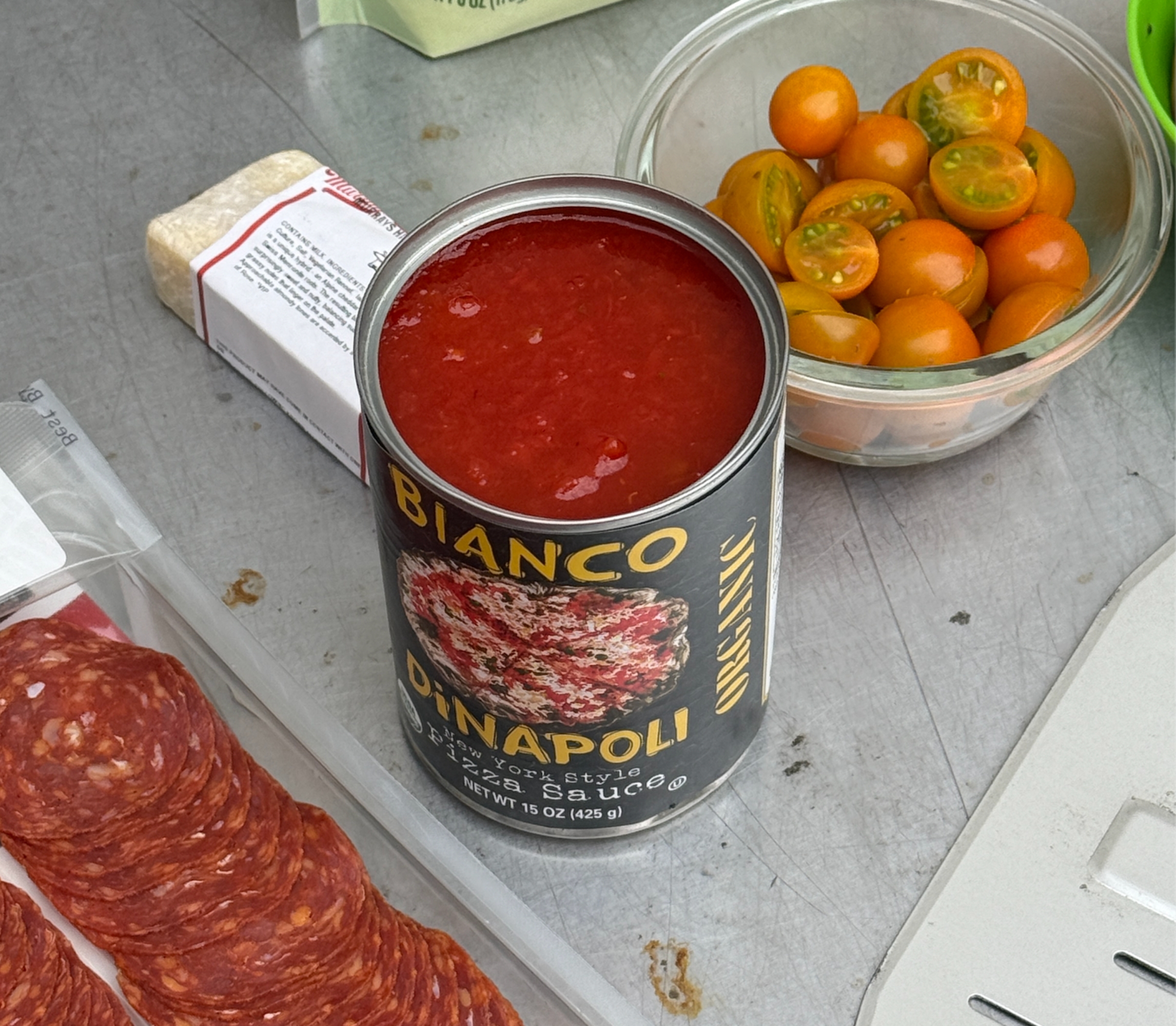Tomatoes, canned tomato sauce, cheese packet, and sliced pepperoni arranged on a table, likely prep for making a dish