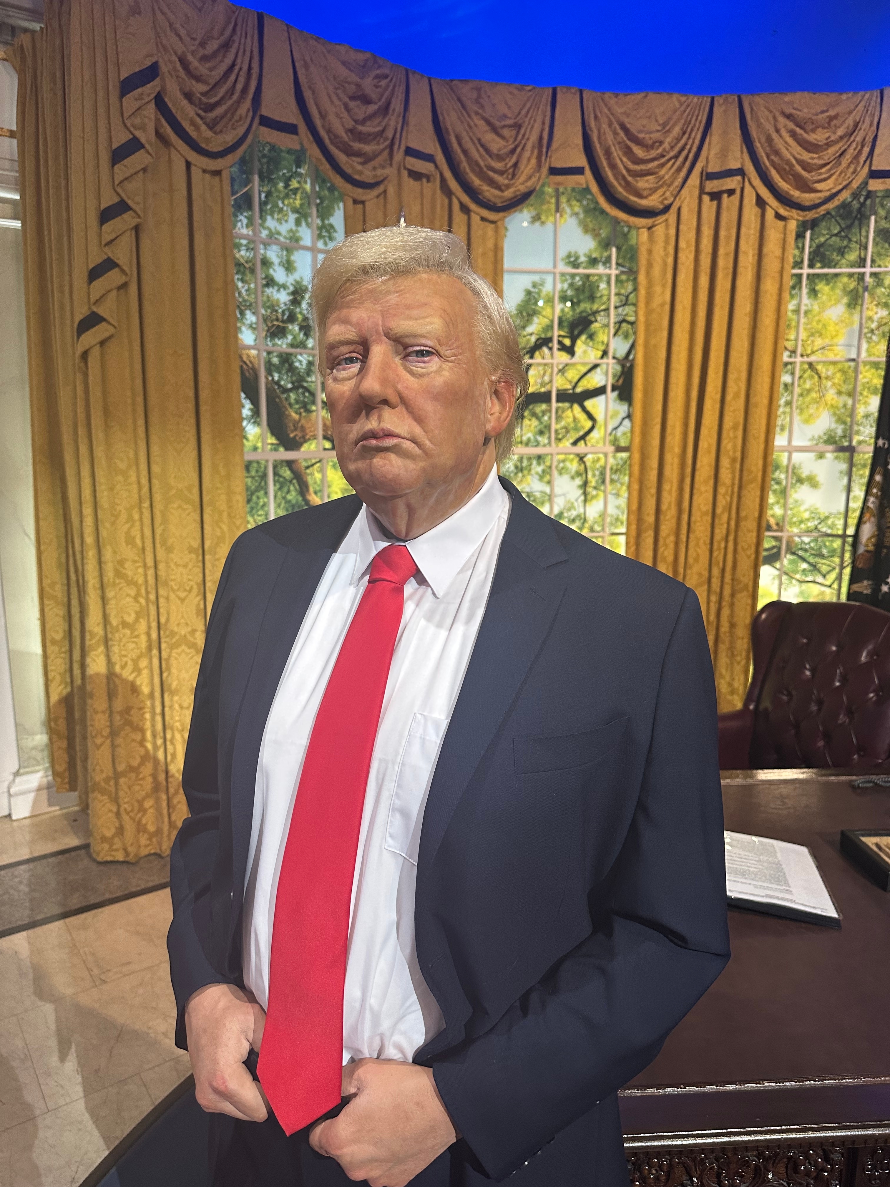 A wax figure of a man in a suit with a red tie stands in an office setting with a large desk and curtain backdrop, evoking a presidential scene