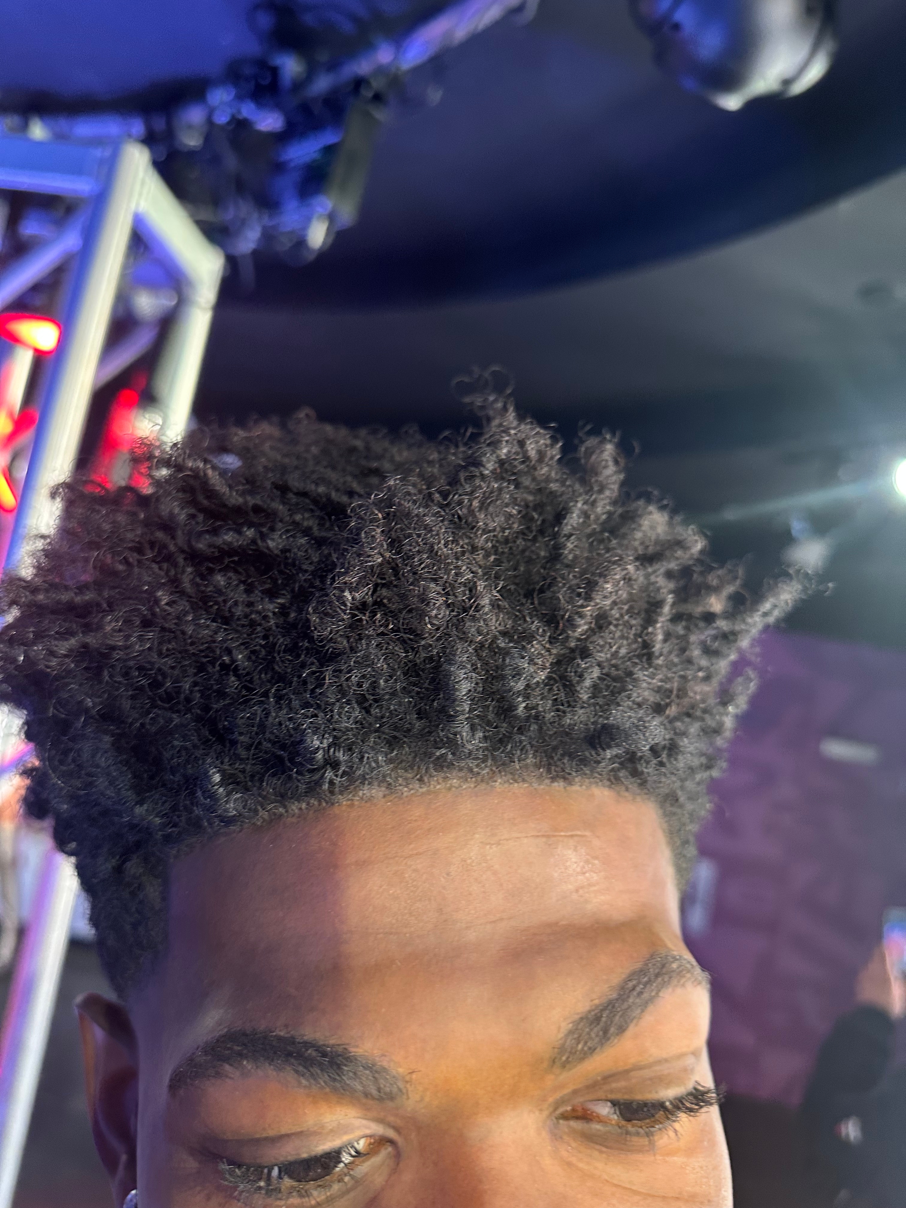 Close-up of a person’s textured hairstyle, against a backdrop of stage lighting and equipment, suggesting a media or event setting