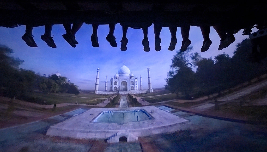 View from a ride with the Taj Mahal in the background and silhouettes of people's legs hanging above, enhancing the sense of height and thrill