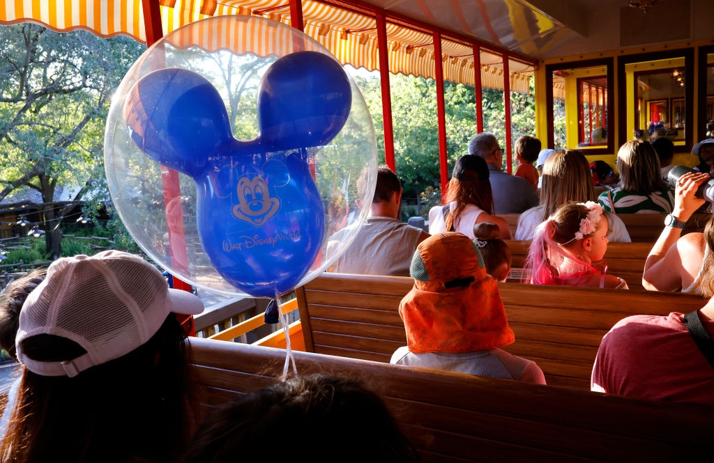Families ride a park train, featuring a Mickey balloon. Children wear hats, some looking outward. A joyful, leisure atmosphere is present