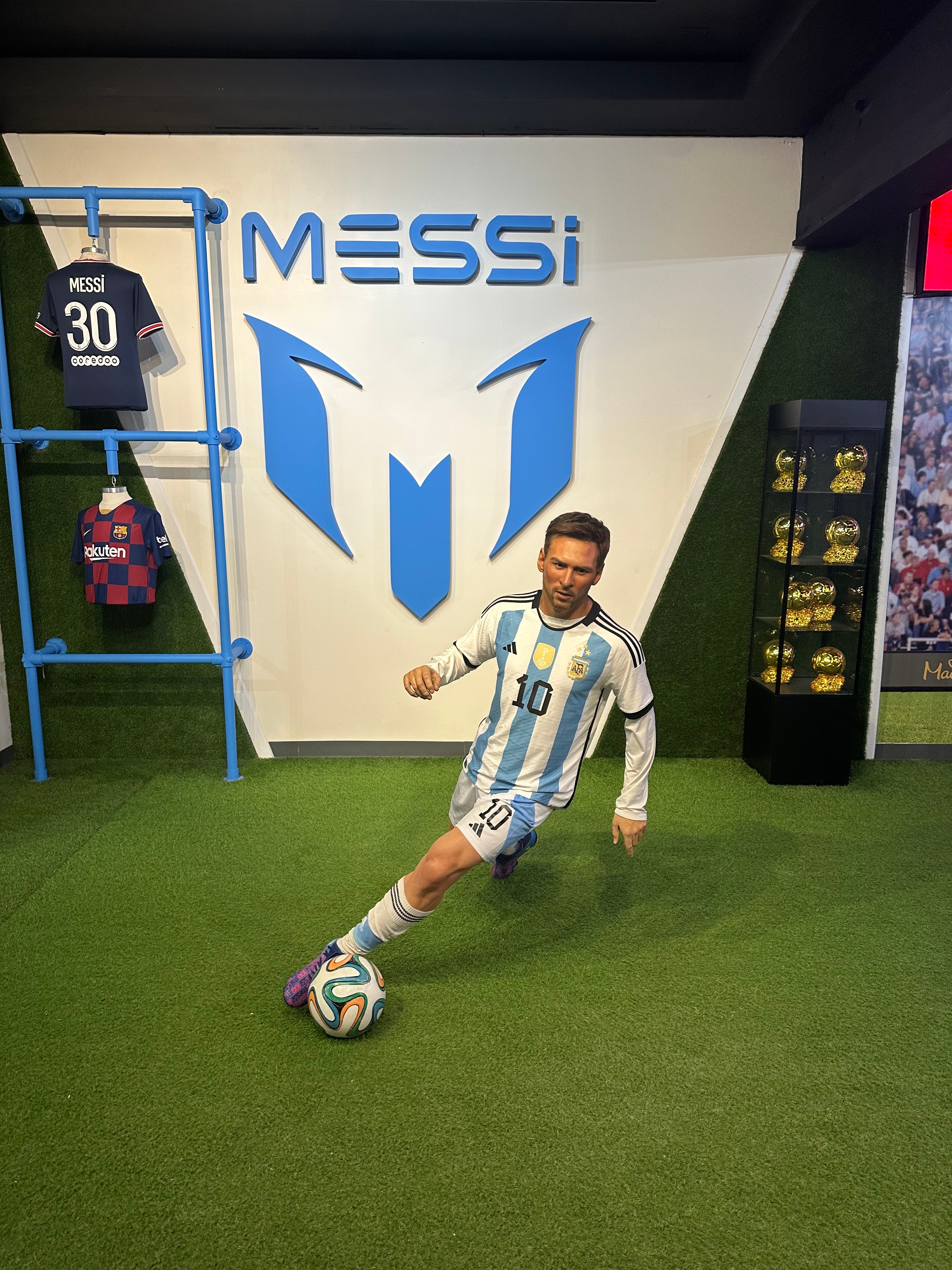 Wax figure of a soccer player in a blue and white uniform, posing with a ball indoors near jerseys and trophies, featured in a travel article