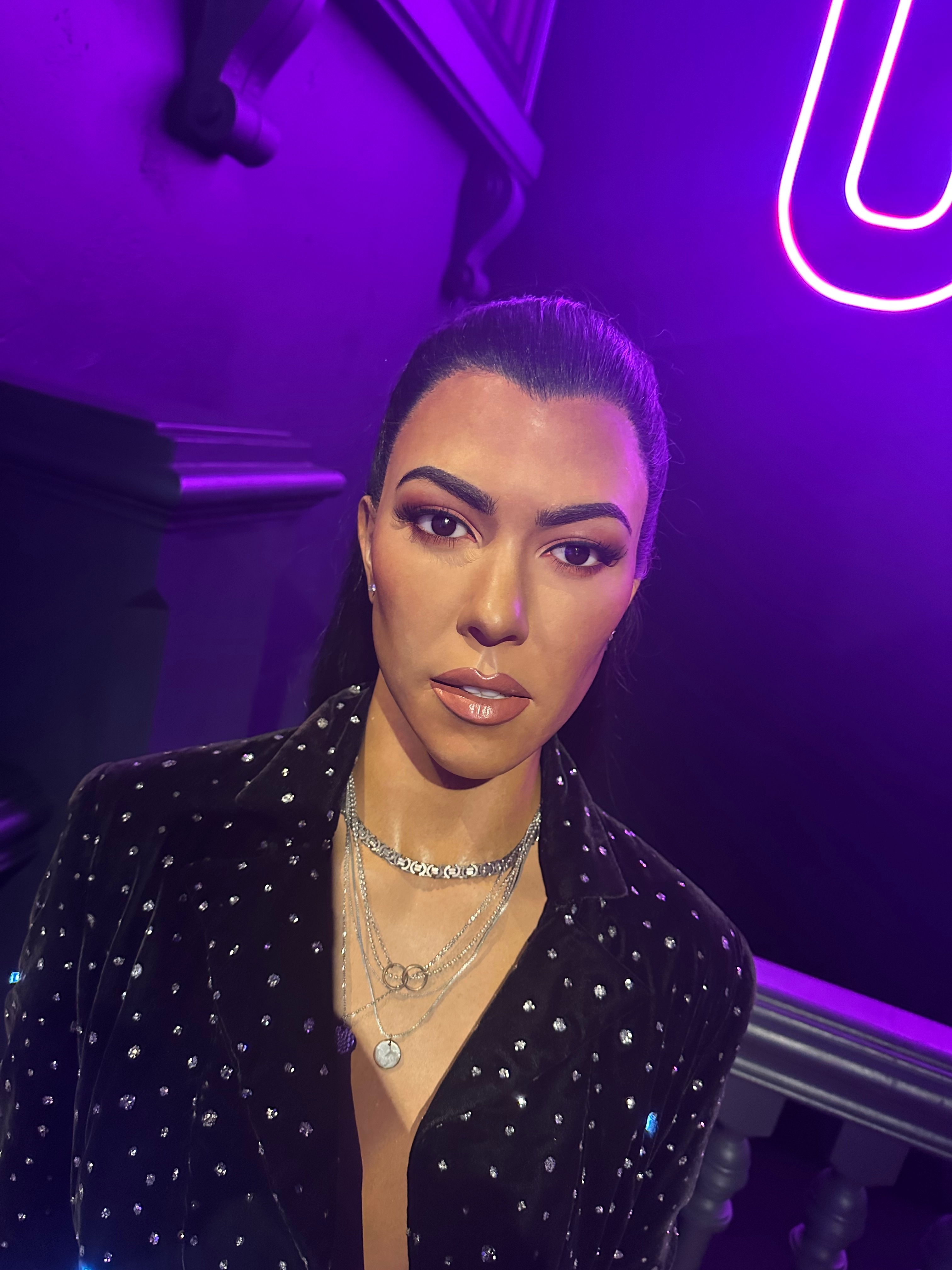 Person in a sparkling, studded jacket with layered necklaces, standing in front of neon lights and railing, suitable for travel article