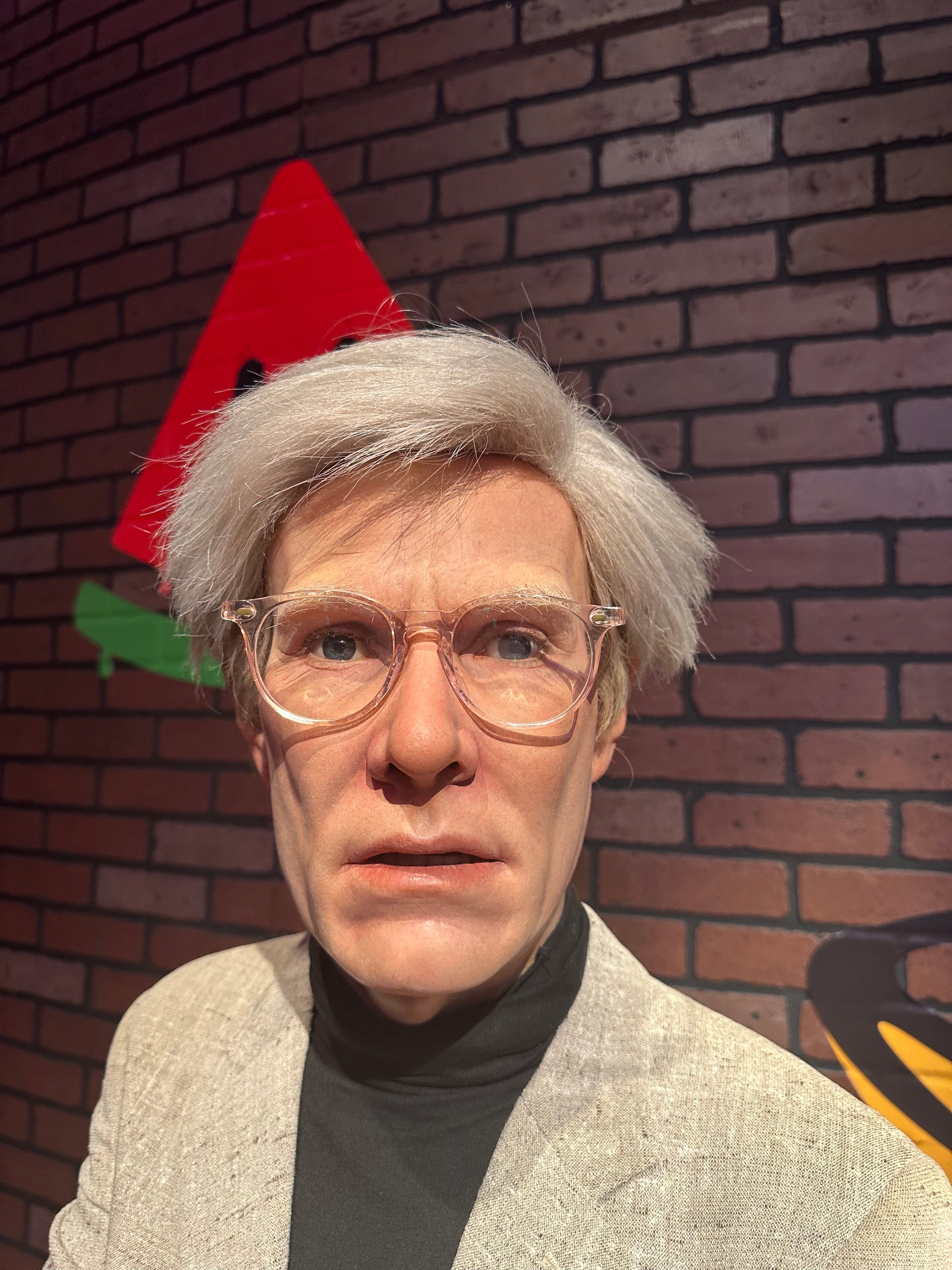 Person in a turtleneck and blazer stands against a brick wall background. The setting suggests an art or celebrity-themed exhibit