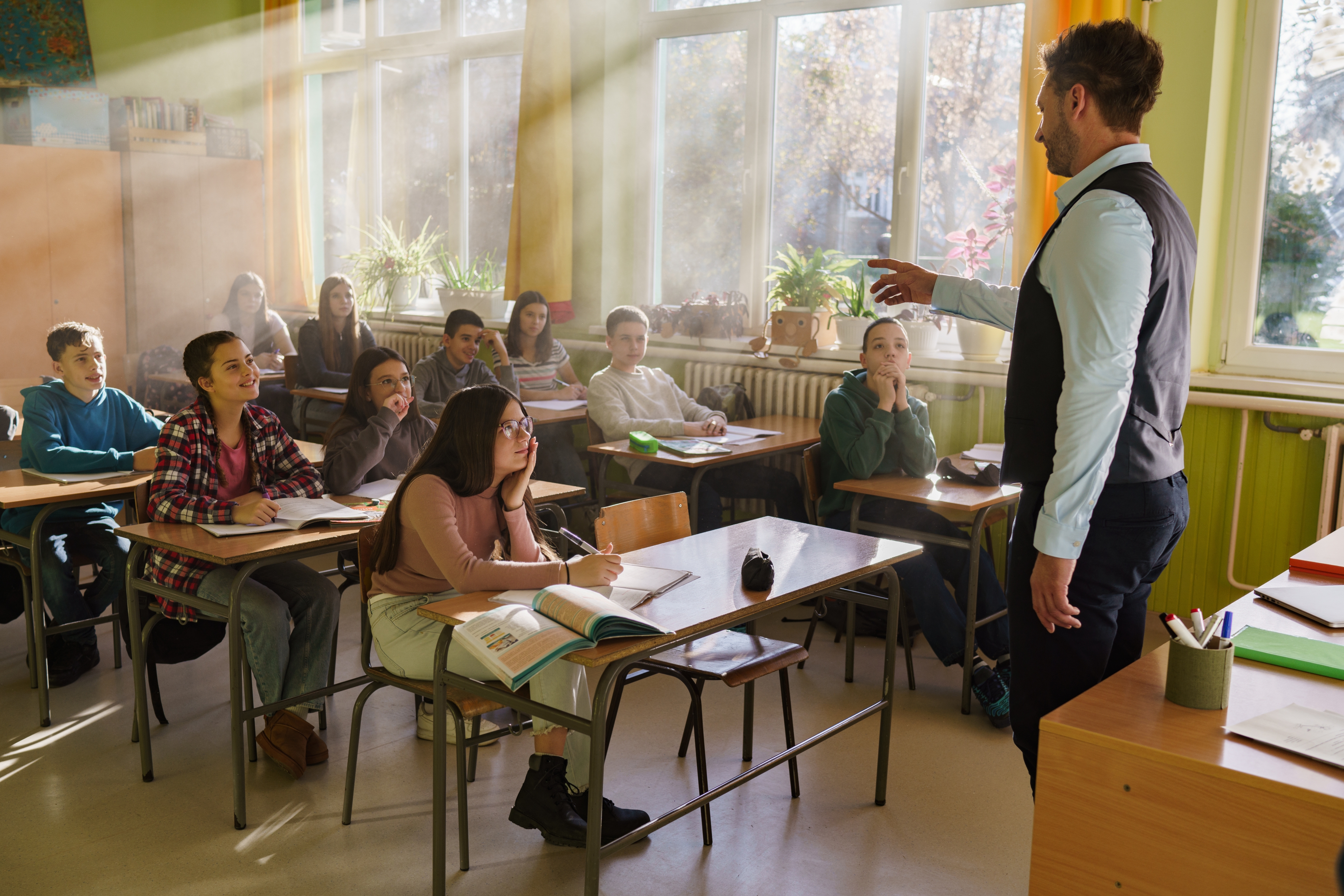 A teacher engages high school students sitting at desks in a well-lit classroom with windows