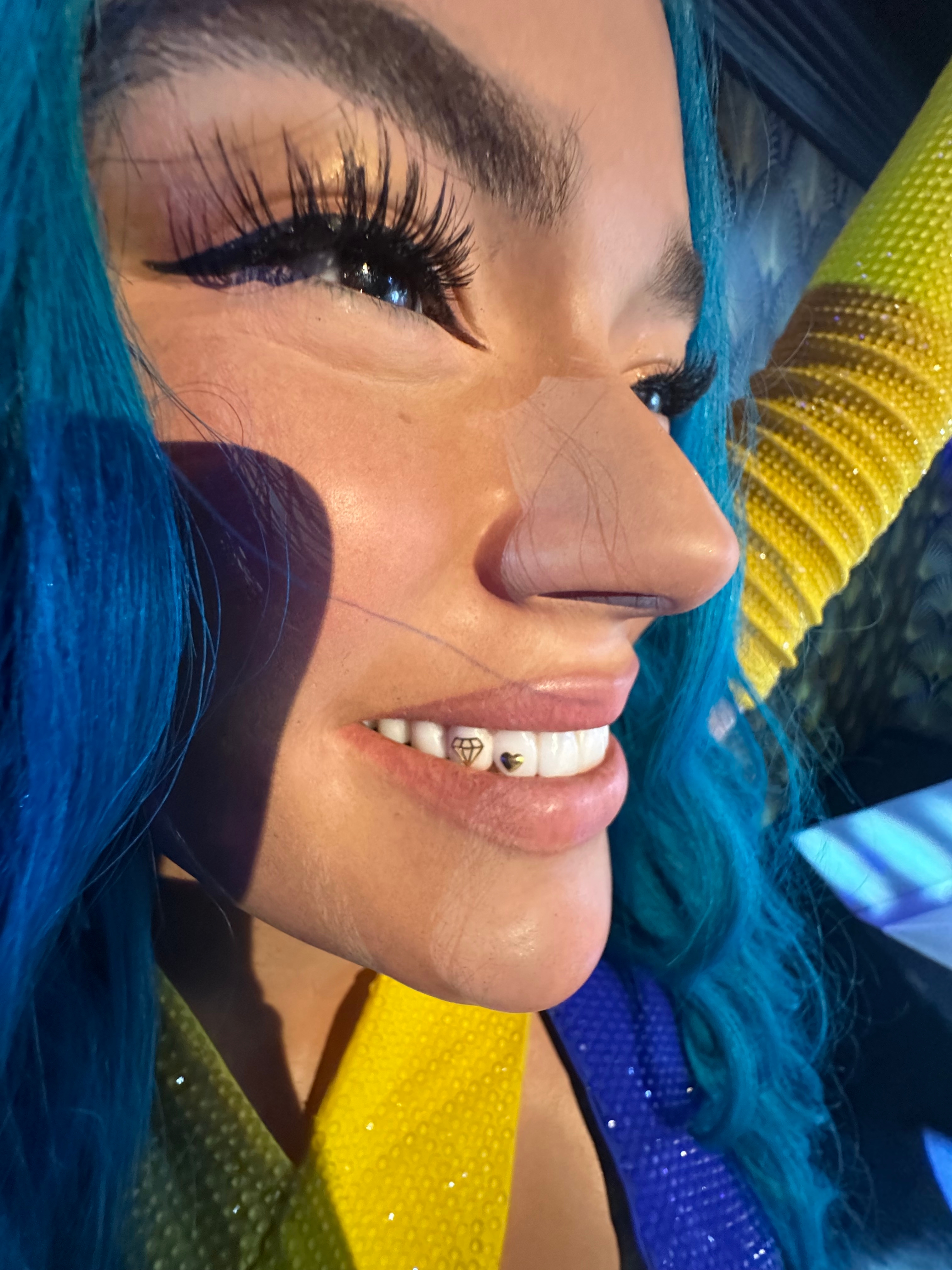 Person with long lashes and colorful braided hair smiles, showing dental jewels. They're wearing a vibrant outfit, suggesting a lively, fun setting