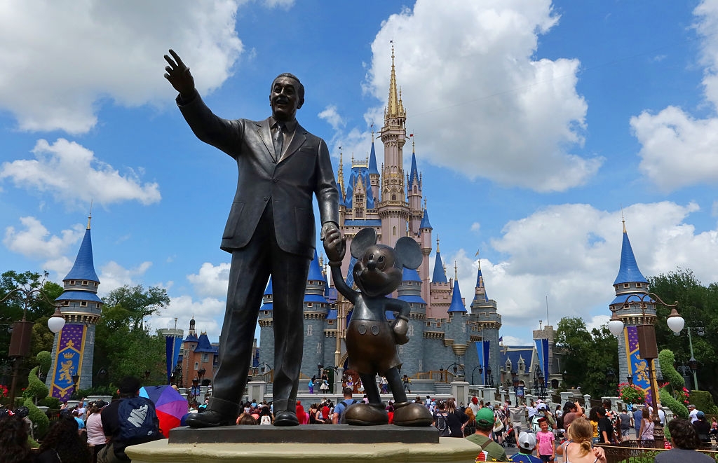 Statue of Walt Disney holding Mickey Mouse's hand in front of a castle at a theme park, with visitors walking around