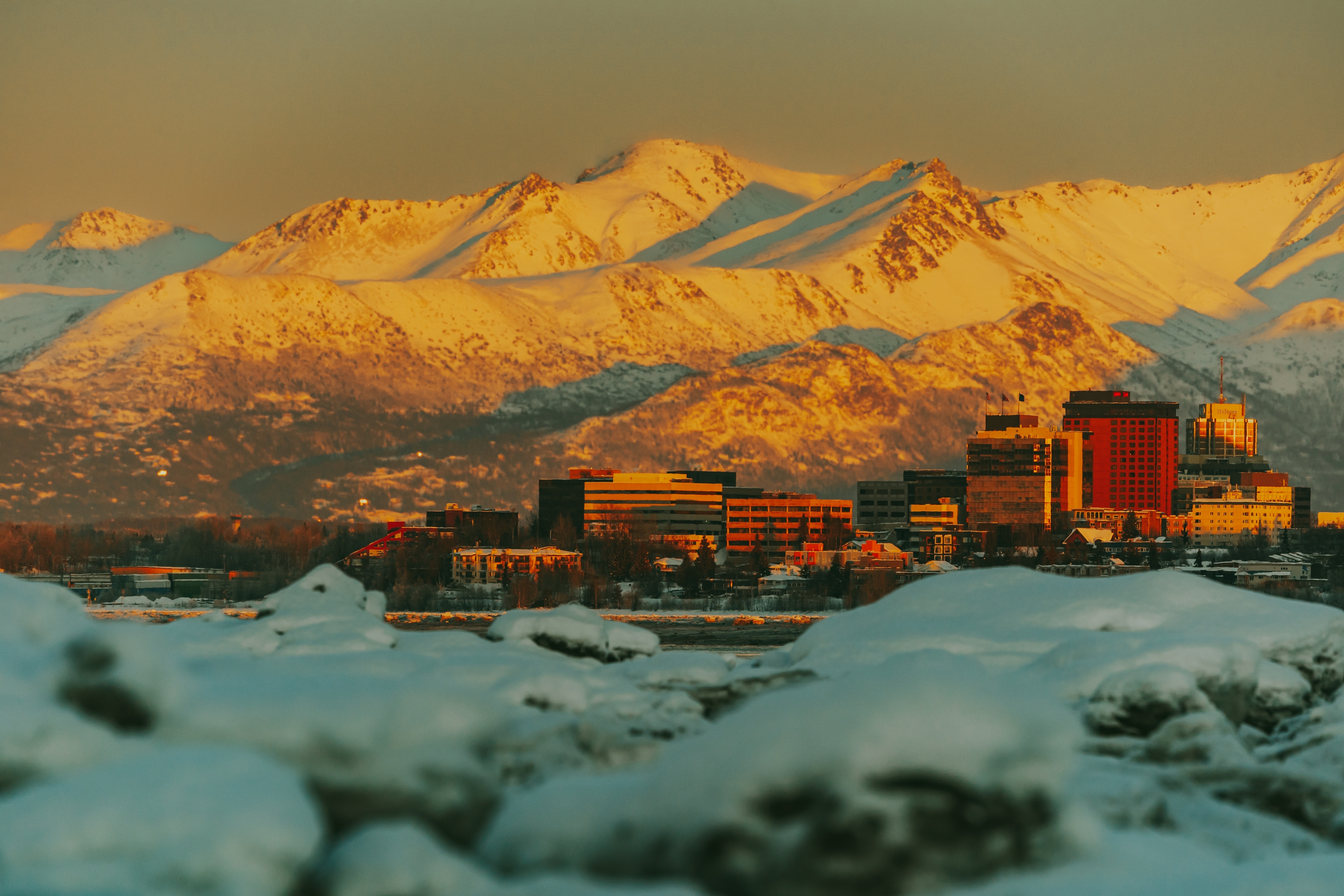 Snowy mountains backdrop a city skyline, capturing a tranquil winter scene with buildings surrounded by snow under a clear sky