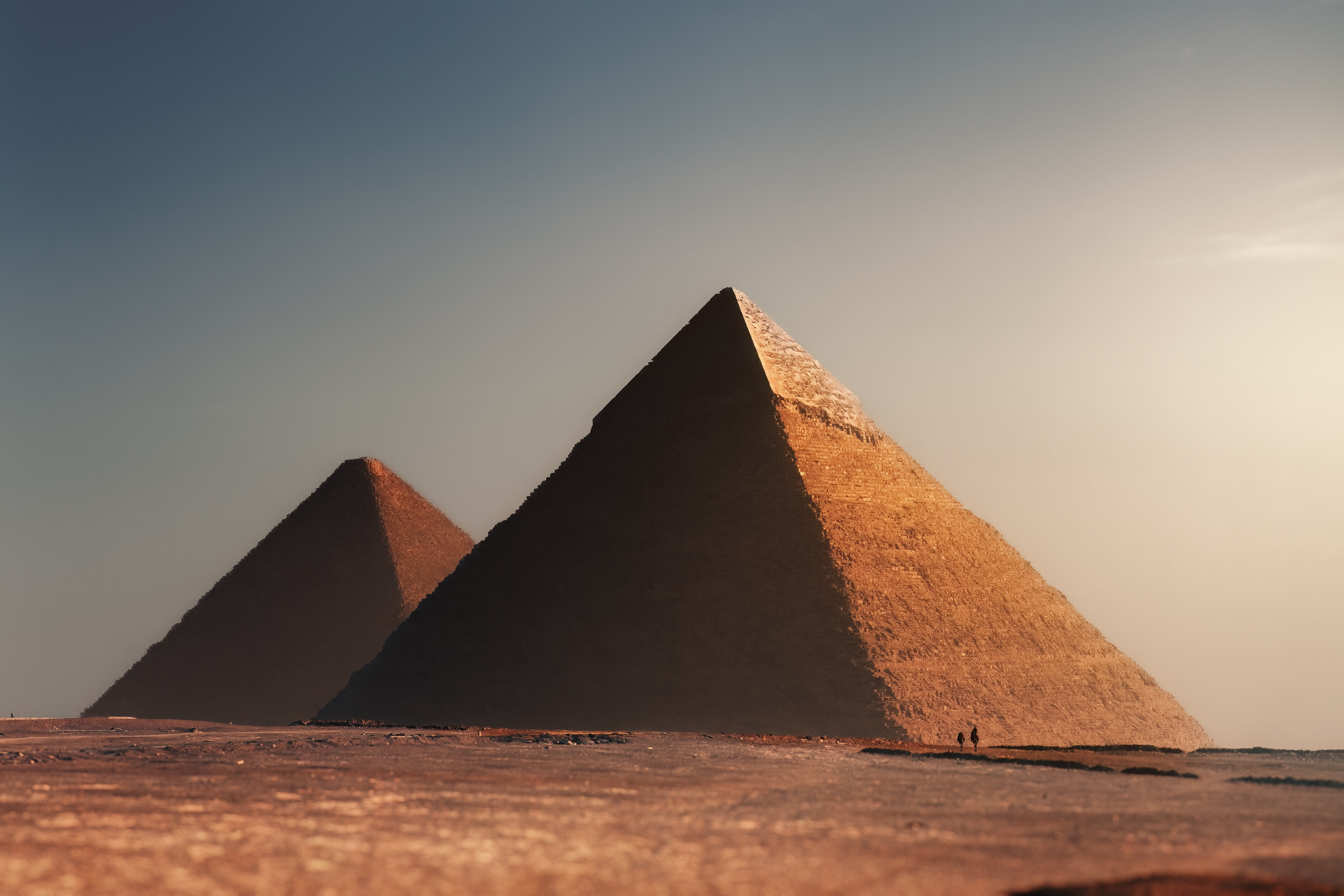 Two large pyramids stand in a desert landscape under a clear sky, with small figures walking nearby, illustrating the vast scale