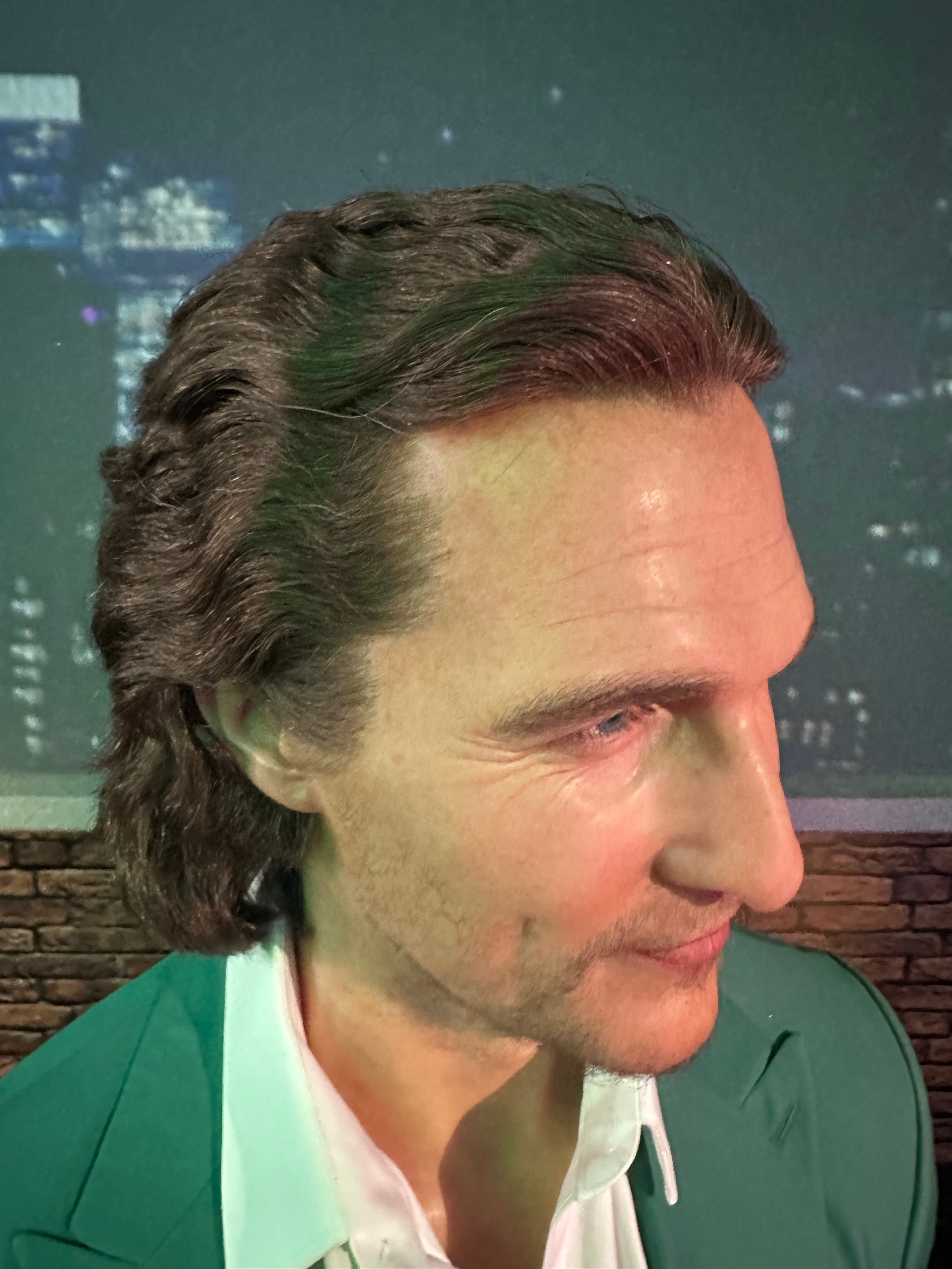 A person with wavy hair in a suit and collared shirt in front of a cityscape backdrop, suggesting a travel or urban setting