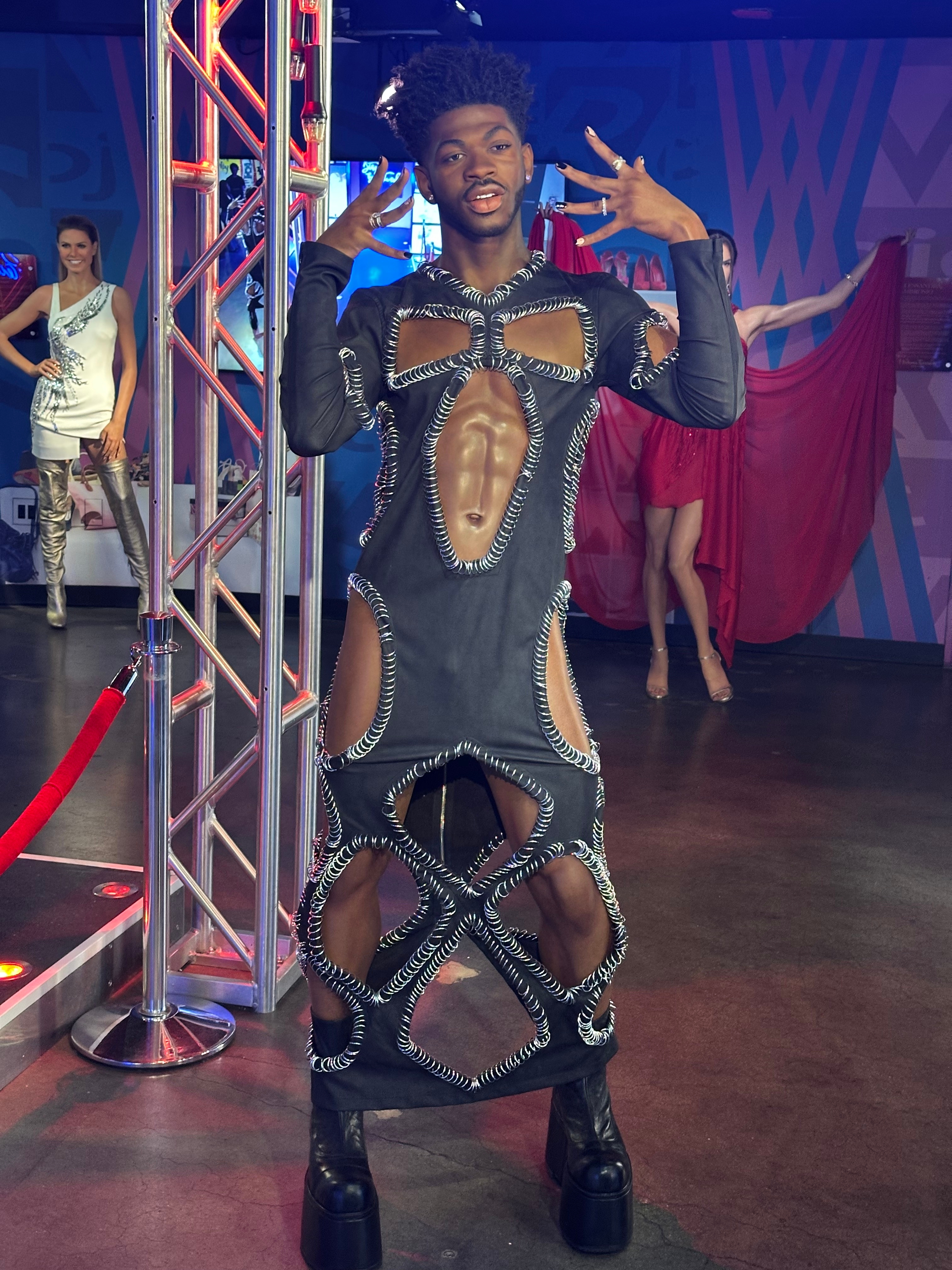 A person on a red carpet poses in an avant-garde dress with cut-out details and high platform shoes, with others in the background