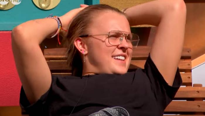A person with clear glasses and a black shirt lounges on a wooden bench, arms behind their head, appearing relaxed and thoughtful