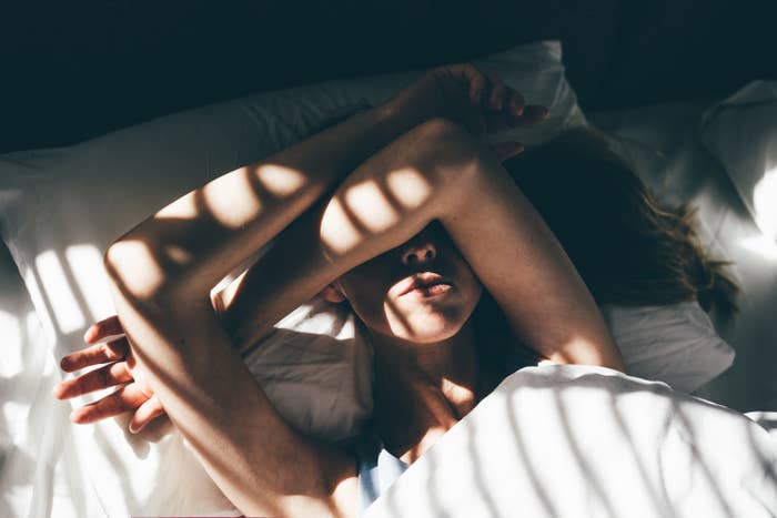 Person lying in bed, blocking sunlight with arms, creating shadow patterns on face; suggests intimacy and morning light