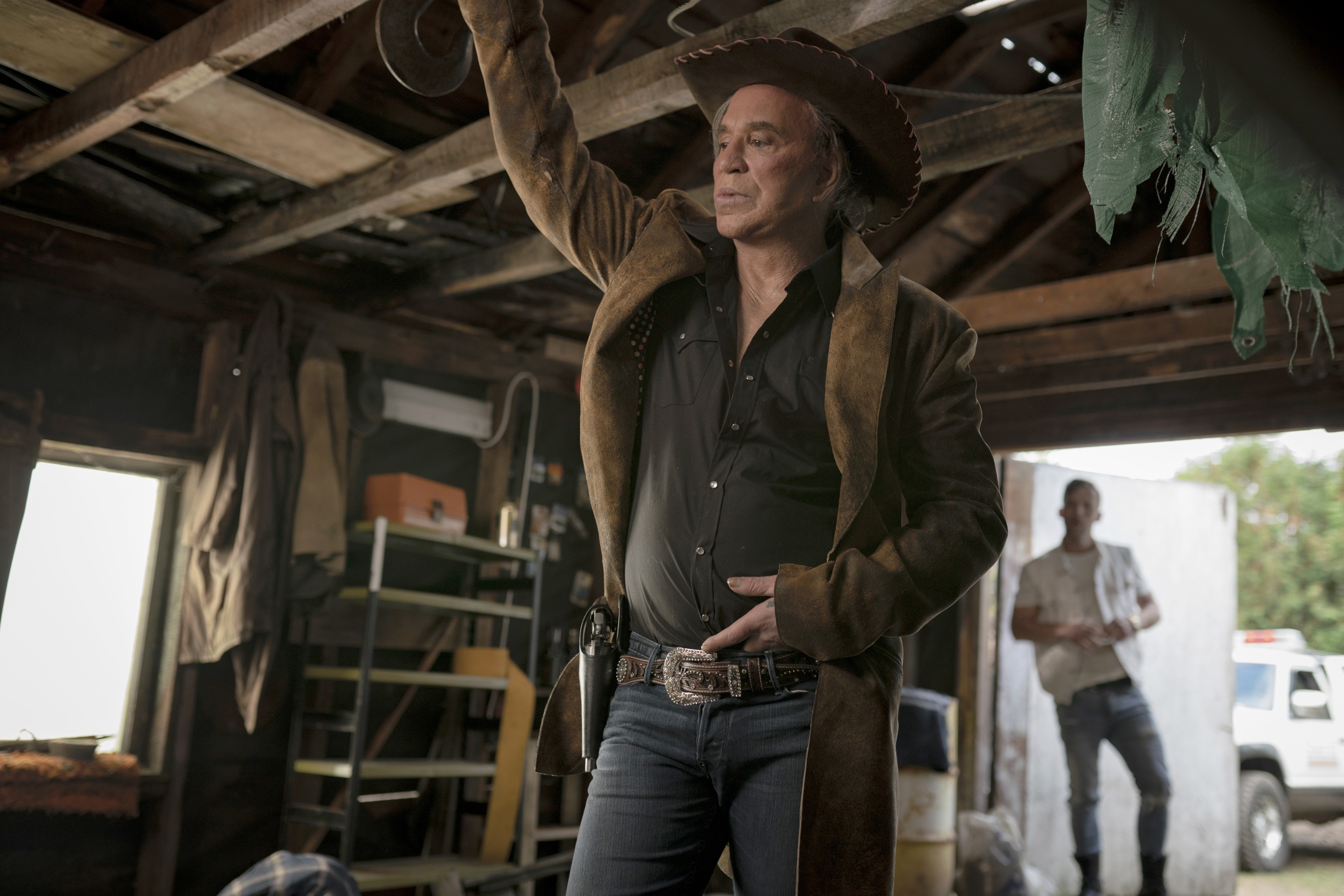 Man in a cowboy hat and long coat stands with hand on hip in a rustic room, exuding a rugged, Western style. Another person stands in the background
