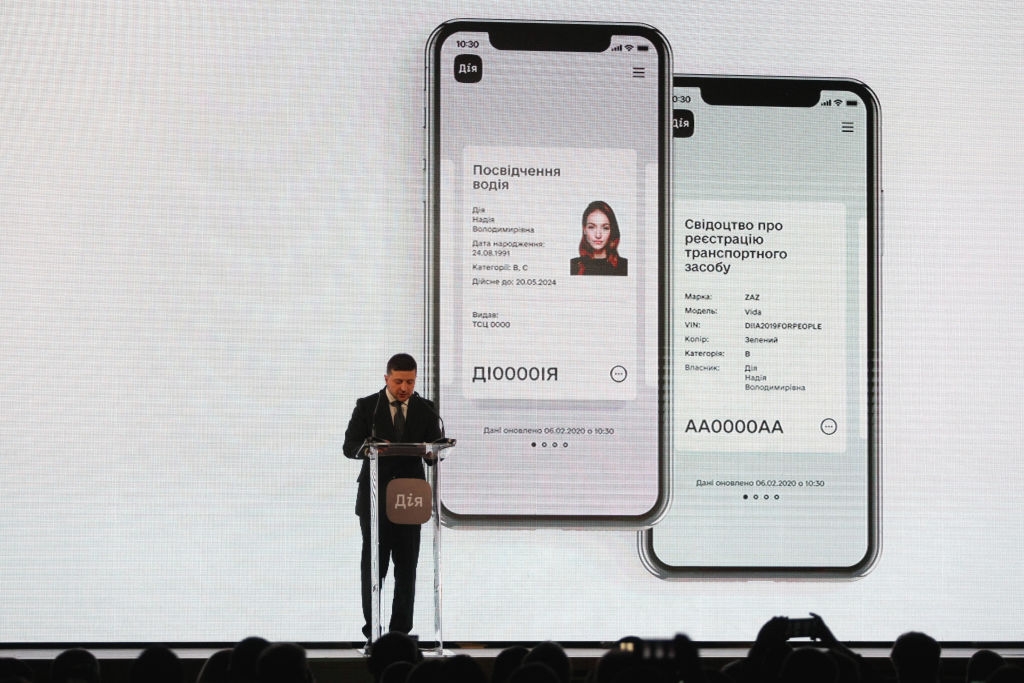 A speaker stands at a podium in front of large screen displays of digital driver's license and vehicle registration on mobile phones