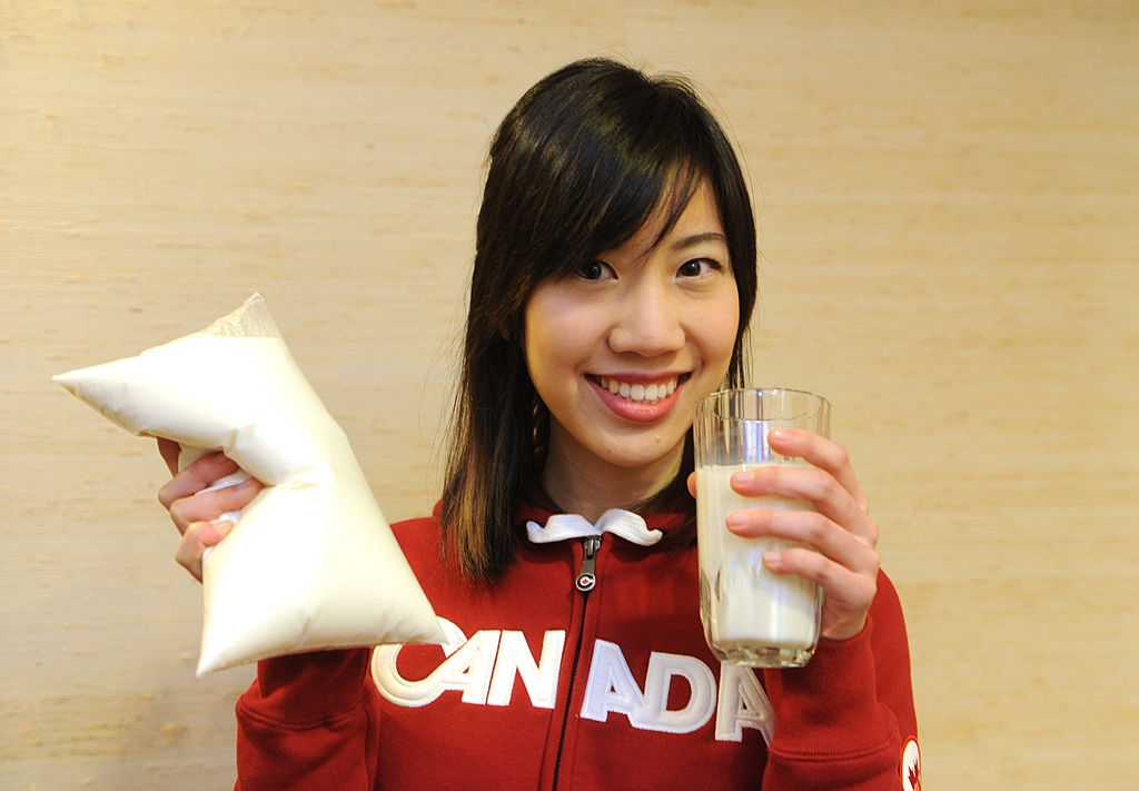 Person smiling, holding a bag of milk in one hand and a glass of milk in the other, wearing a red hoodie with "Canada" written on it