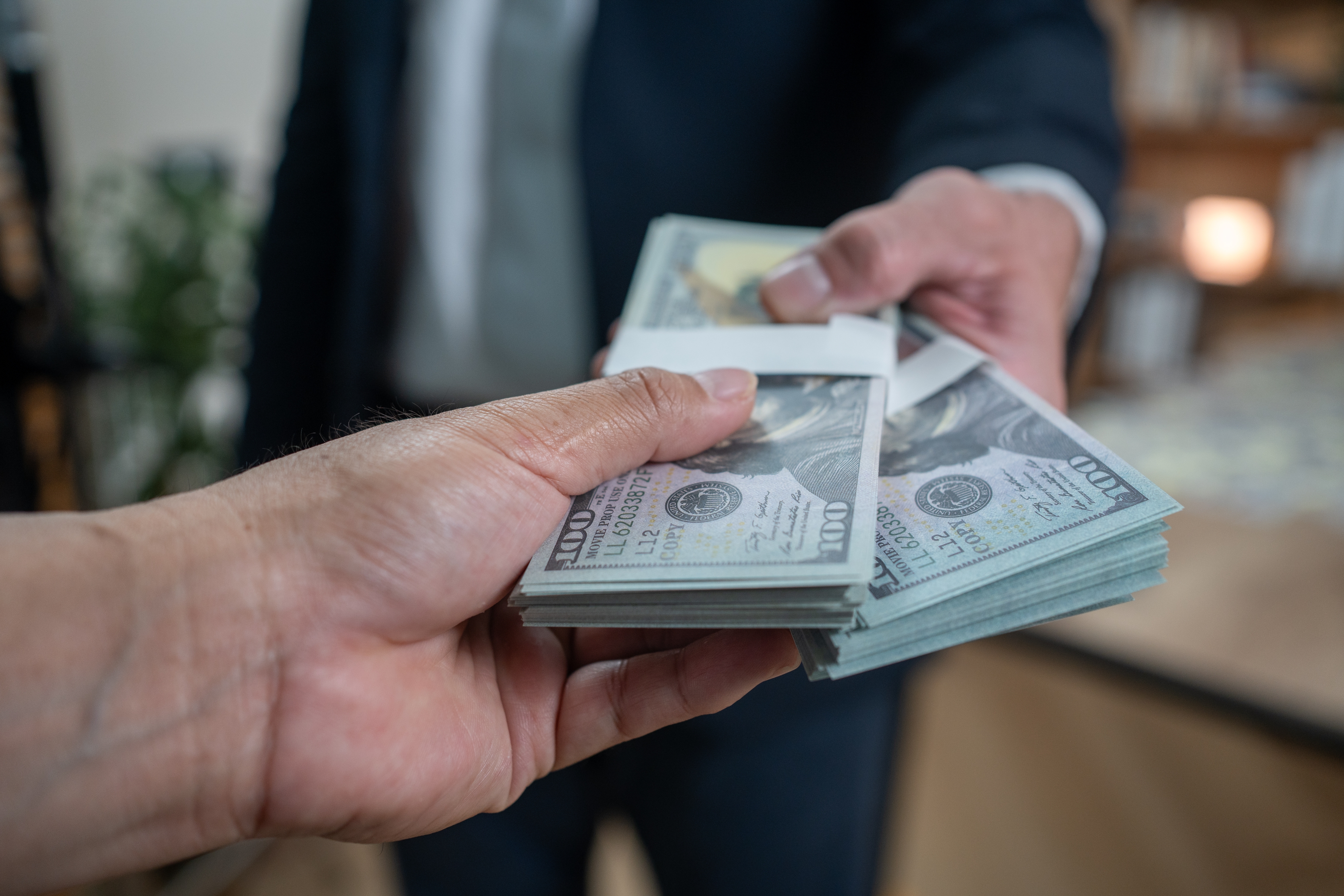 Close-up of a person handing over a stack of US hundred-dollar bills to another person indoors