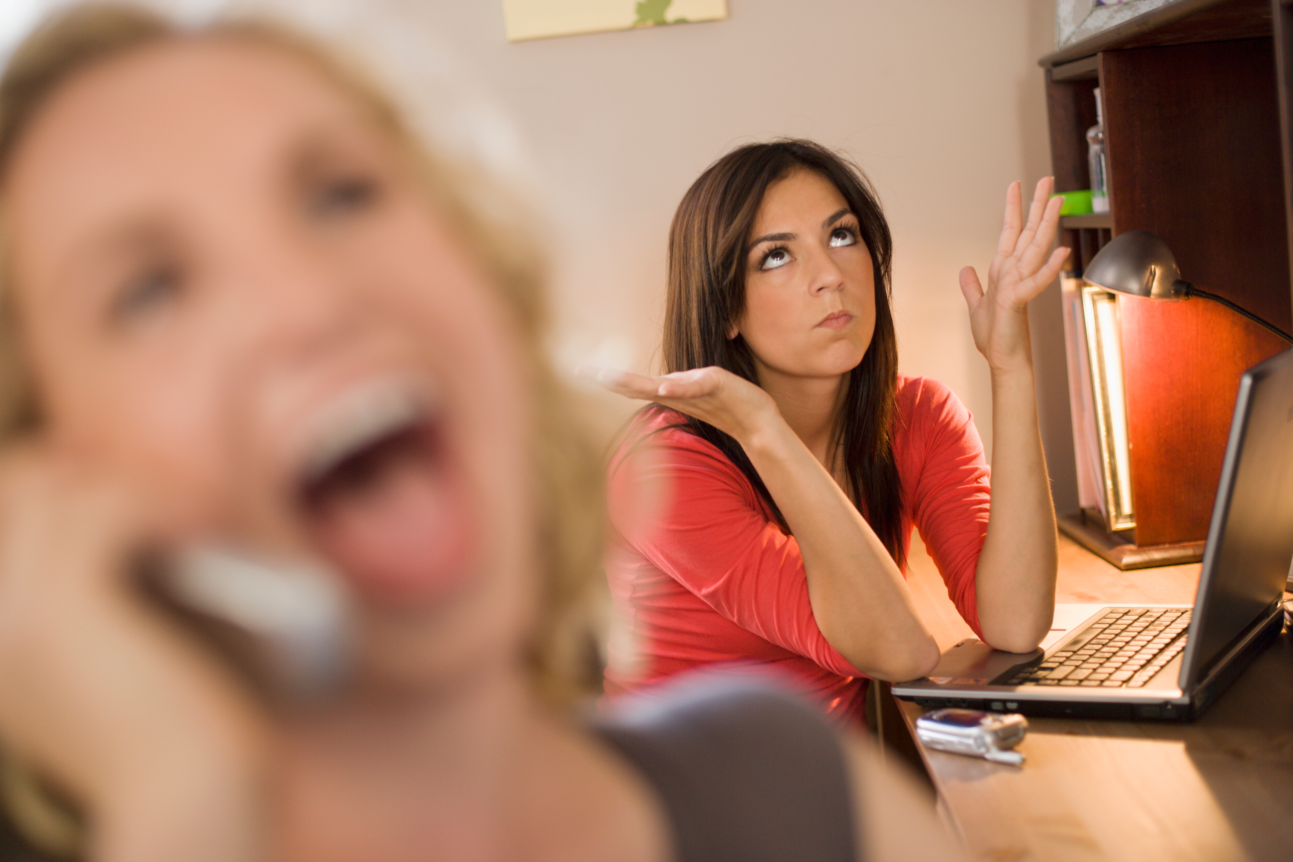 Woman at desk looking frustrated while another person laughs loudly on the phone in the foreground