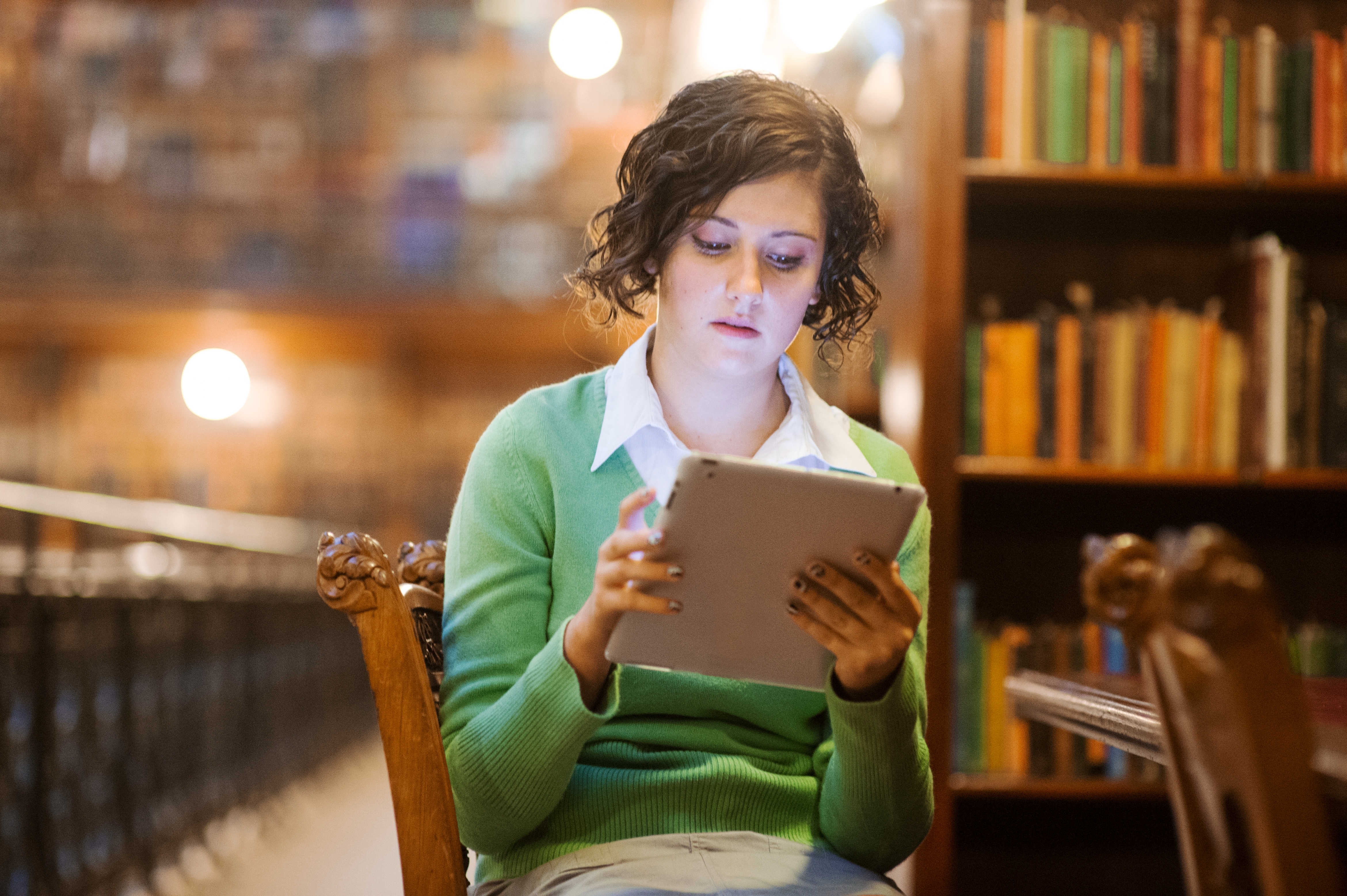 Person reading on a tablet in a library, seated on a wooden chair with books and blurred lights in the background