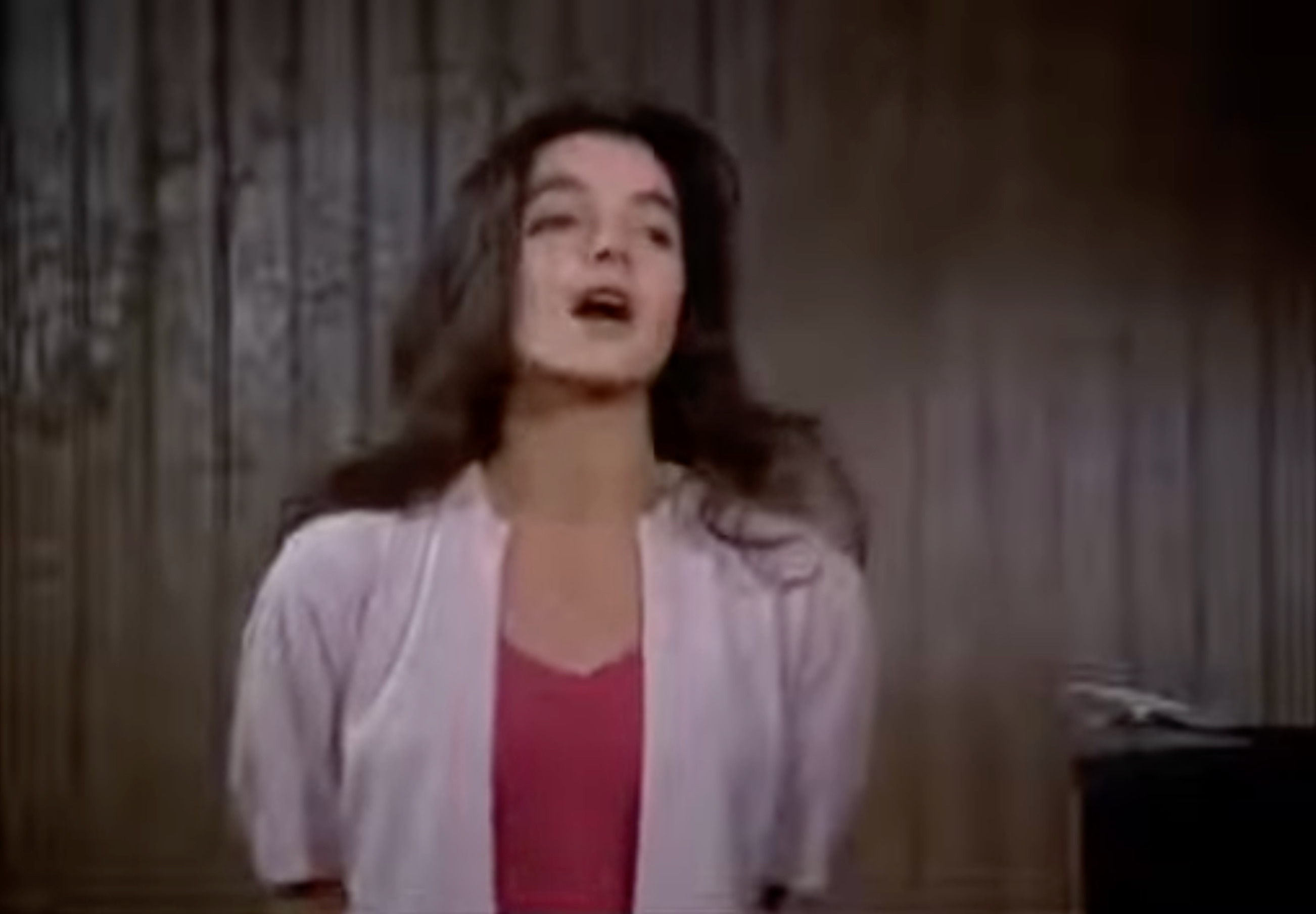 A person sings with their eyes closed, wearing a casual top and a light jacket