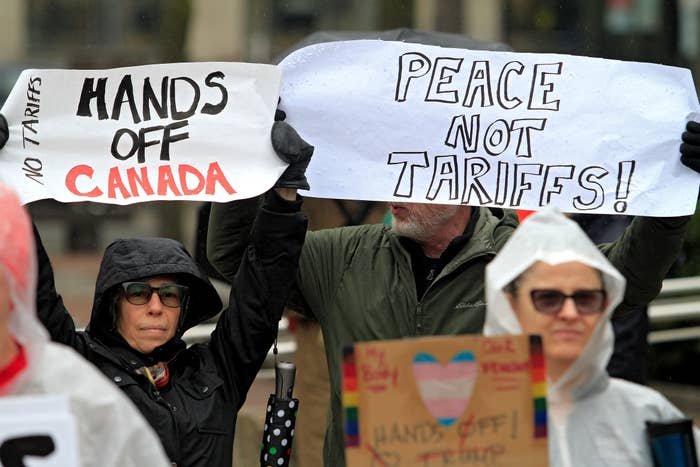 People at a protest hold signs reading "Hands Off Canada" and "Peace Not Tariffs." Protesters wear rain gear