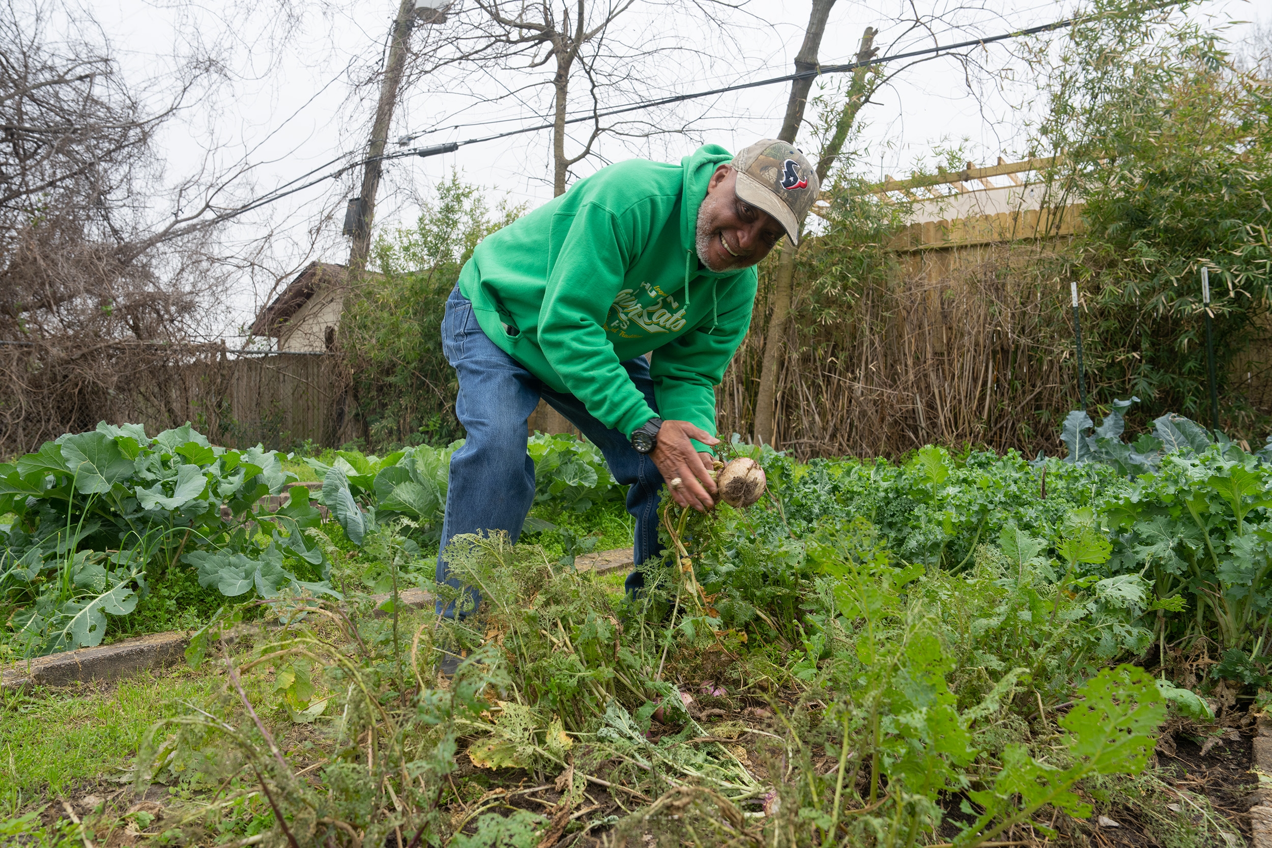 Person in a garden harvesting vegetables, smiling and bending down. Surrounded by lush plants and trees