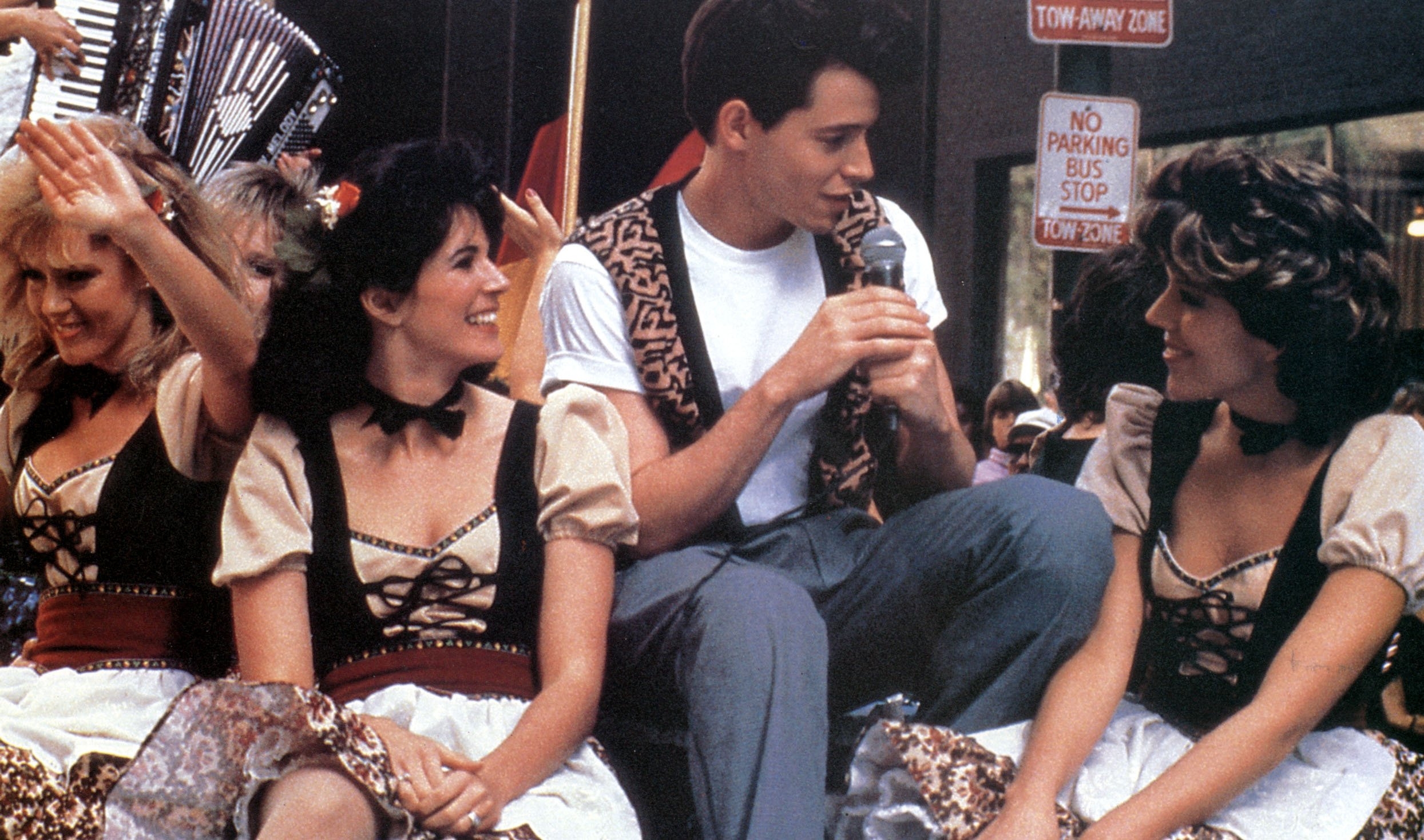 A person in a white T-shirt and patterned vest sings with performers in traditional attire during a cheerful outdoor event