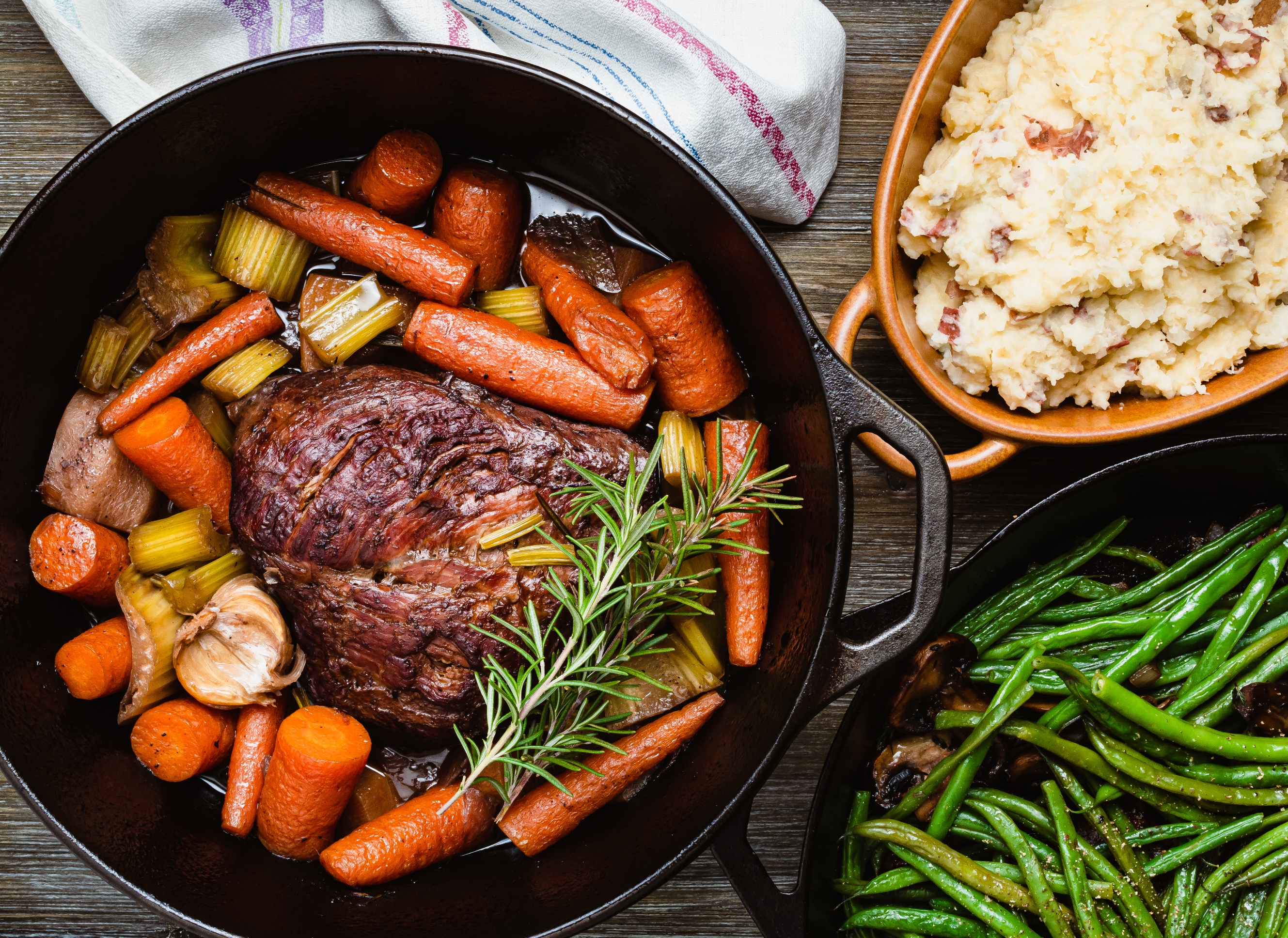 A pot roast in a black dish with carrots, celery, garlic, and fresh herbs. Sides of mashed potatoes and green beans are next to it