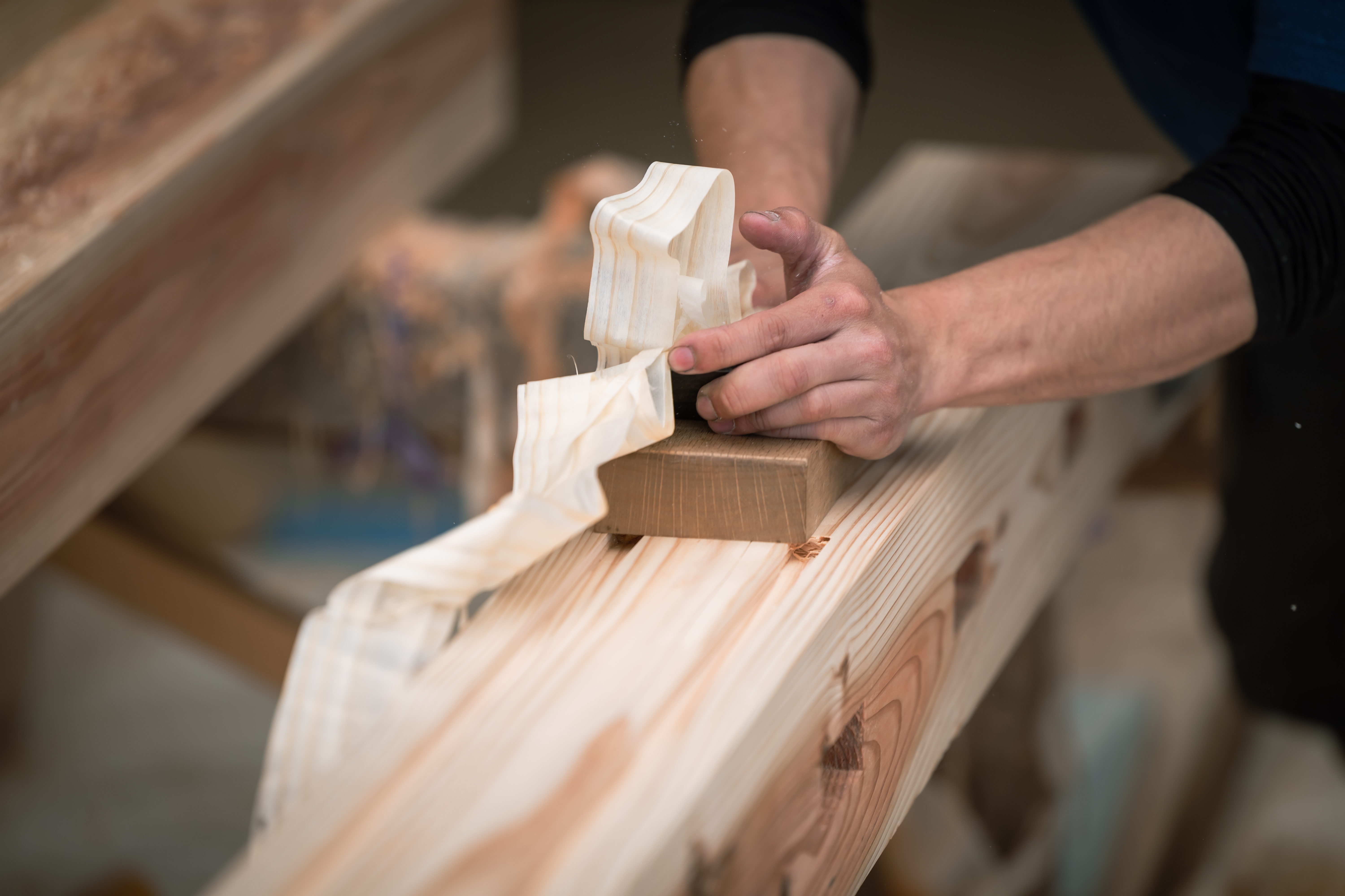 Person using a wood planer to smooth a wooden beam, with shavings curling away