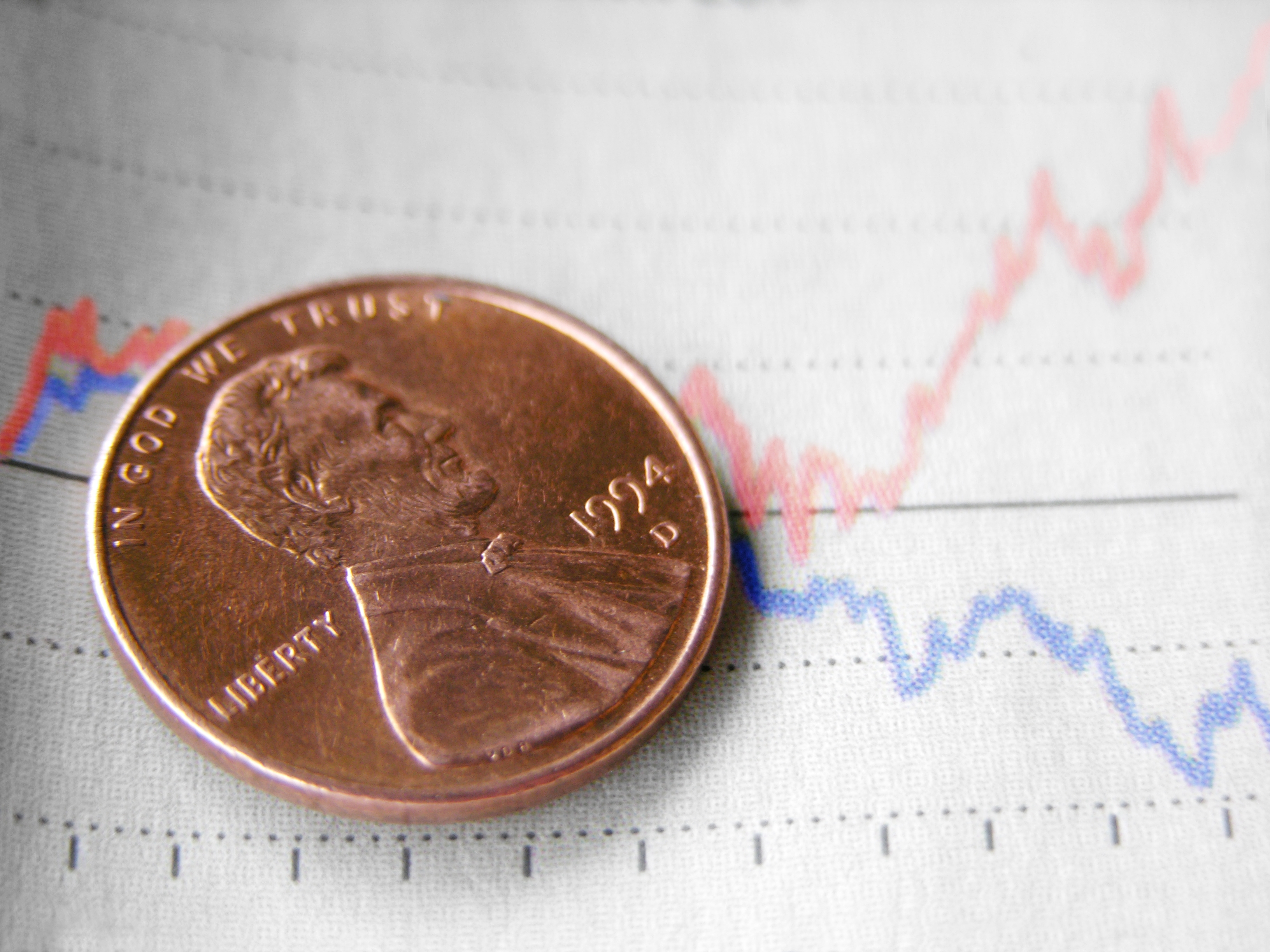 A penny dated 1994 rests on a paper with fluctuating red and blue line graphs, suggesting financial data or stock market trends