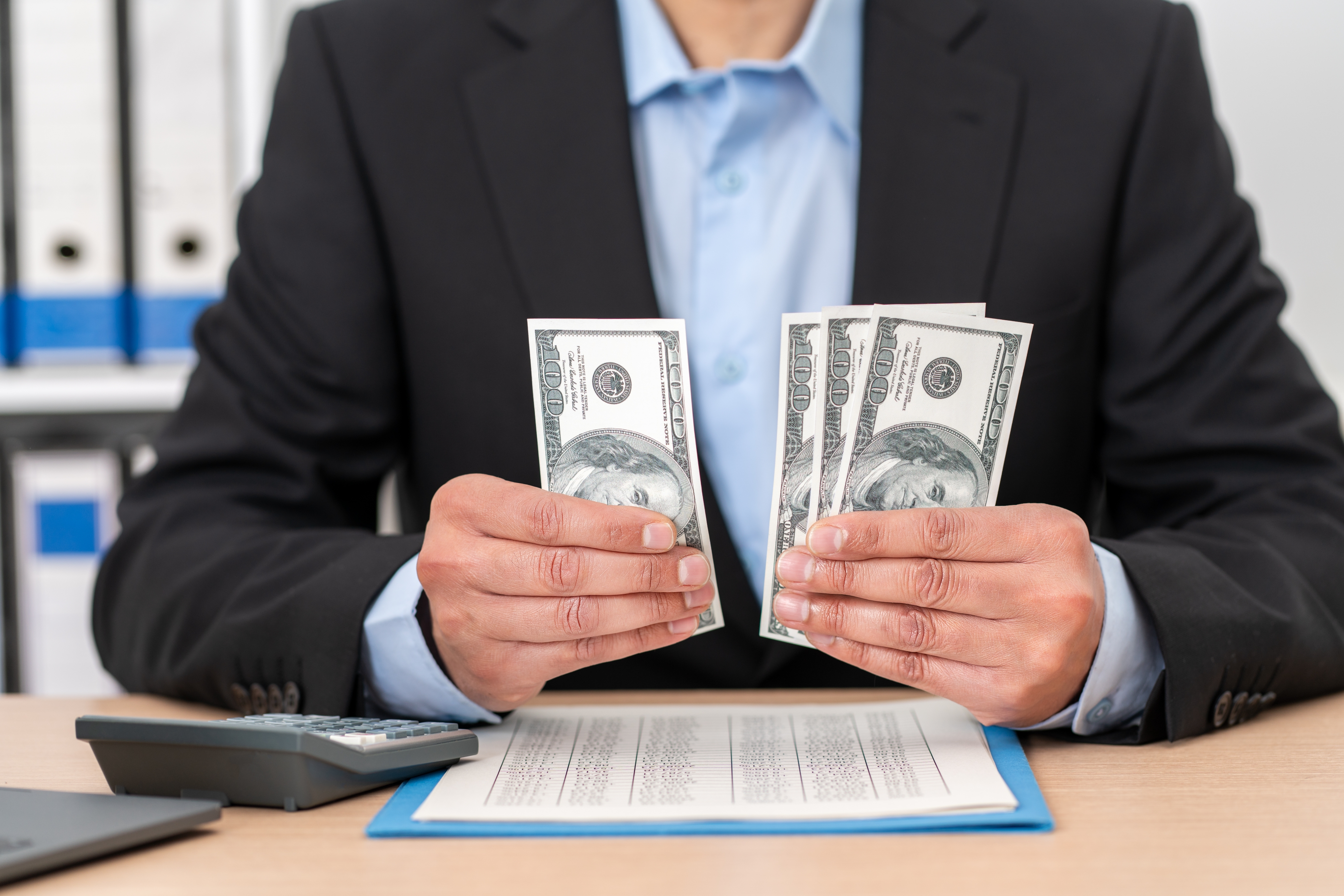 Person in suit counting money at a desk, with a calculator beside a document