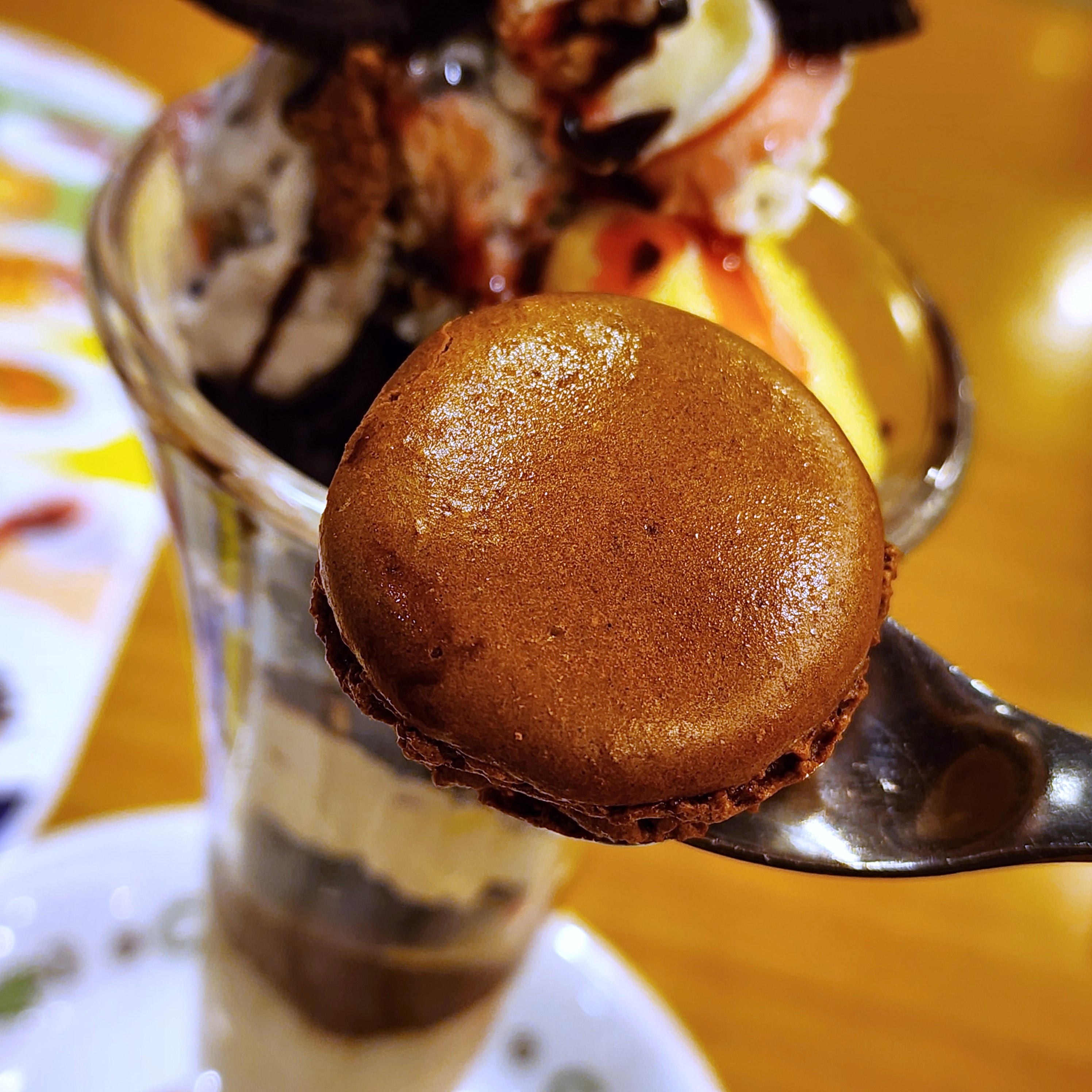 チョコレートマカロンが載ったパフェがスプーンに持ち上げられている。背景にはメニューがぼんやり見える。