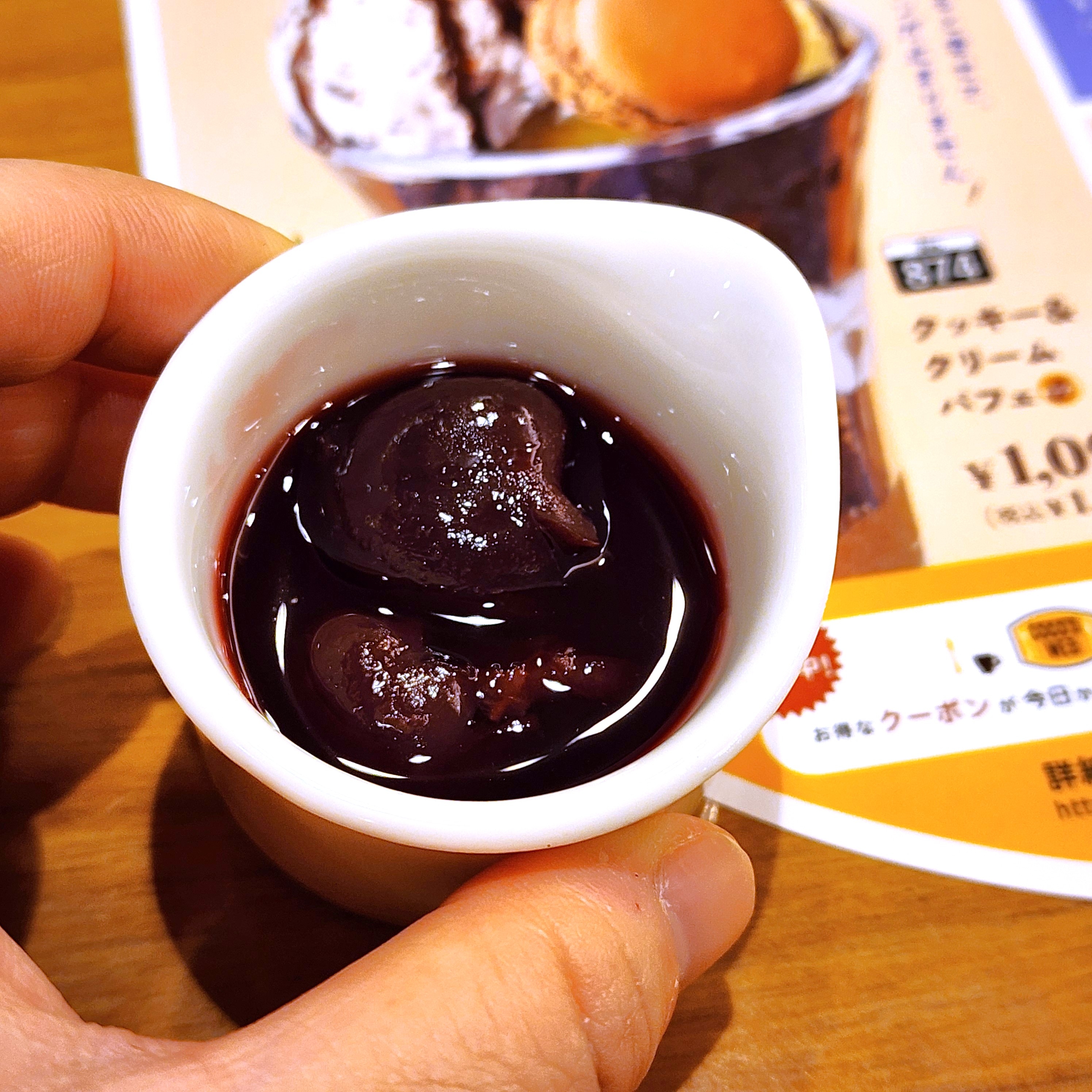 小さなカップに入ったクリーム状のデザートが、人の手で持たれている。背景に広告の一部が見える。