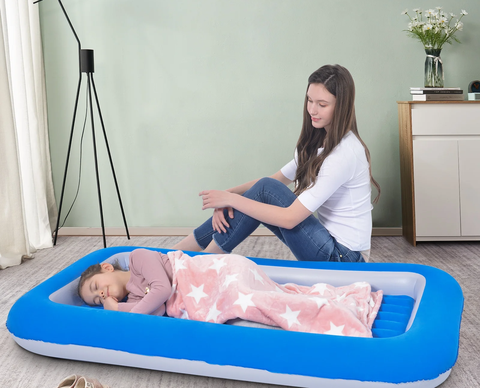 Child sitting beside a child sleeping on an inflatable toddler bed with a star-patterned blanket, in a living room