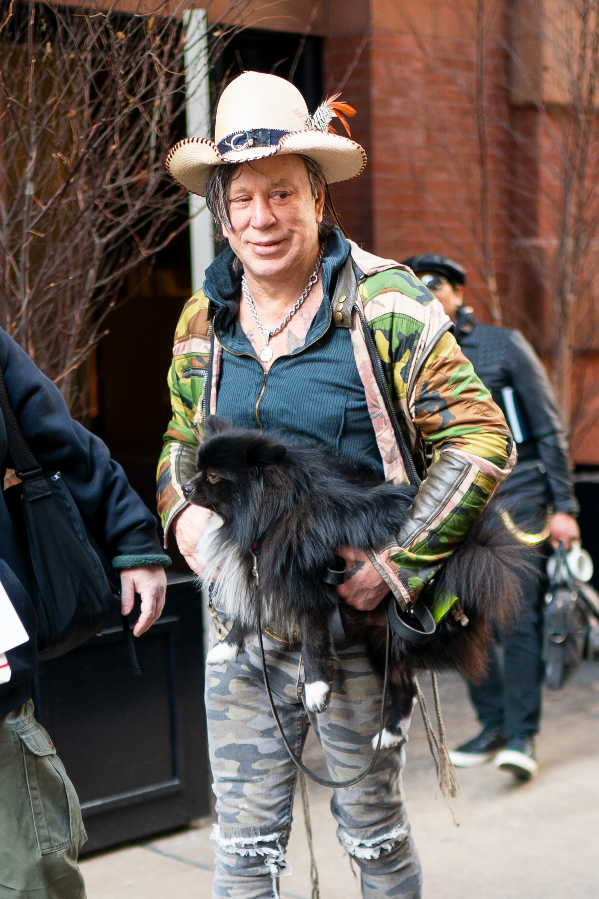 Mickey Rourke in a patterned jacket, cowboy hat with feathers, and camo pants holds a fluffy black dog