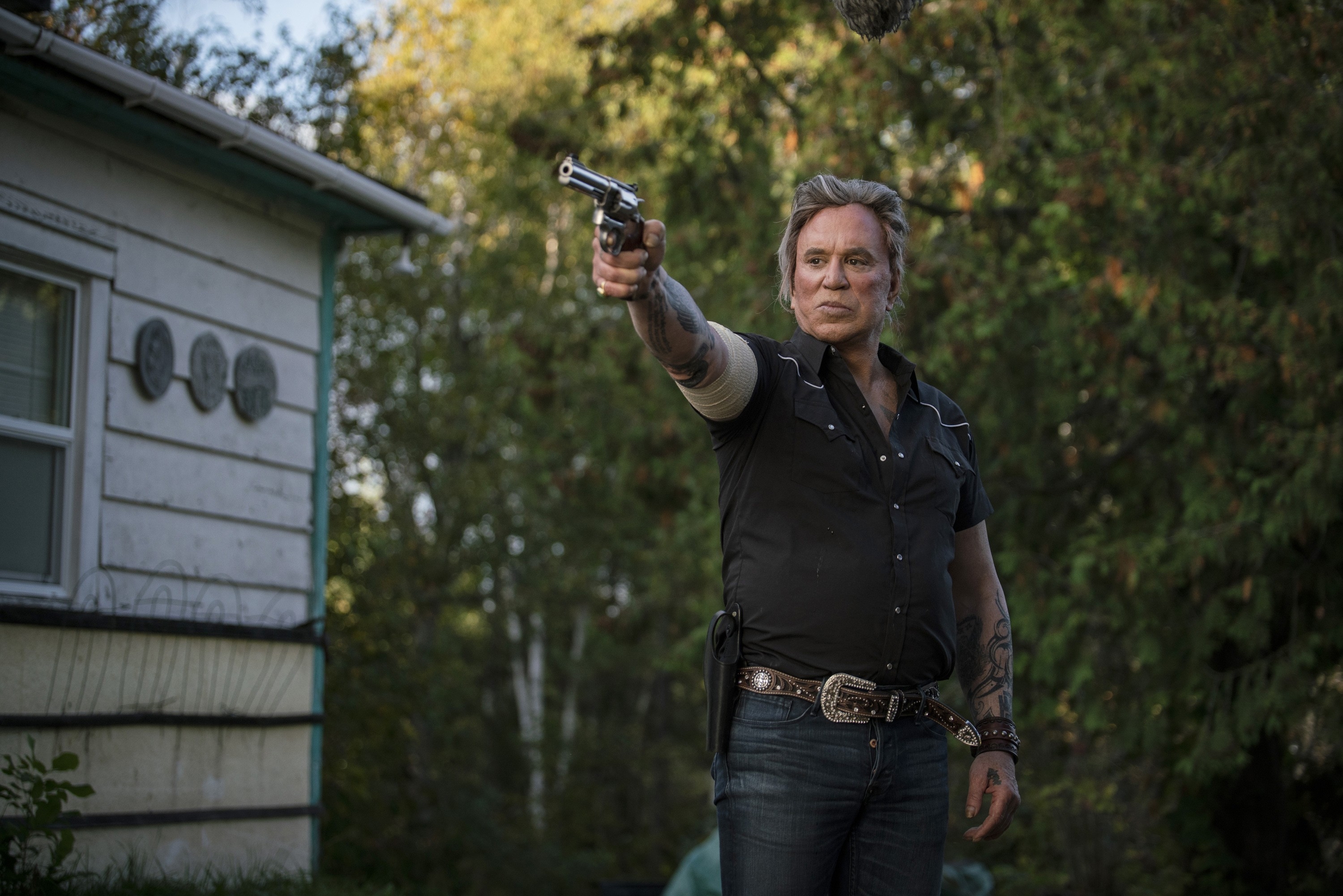 Mickey Rourke outdoors pointing a gun, wearing a short-sleeve shirt, jeans, and a belt with a large buckle. Trees and a house visible in the background