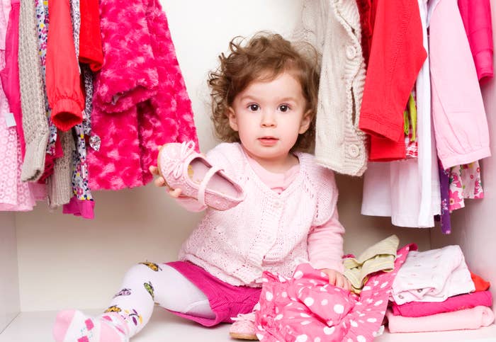 Child in a wardrobe holds a shoe, surrounded by various clothes