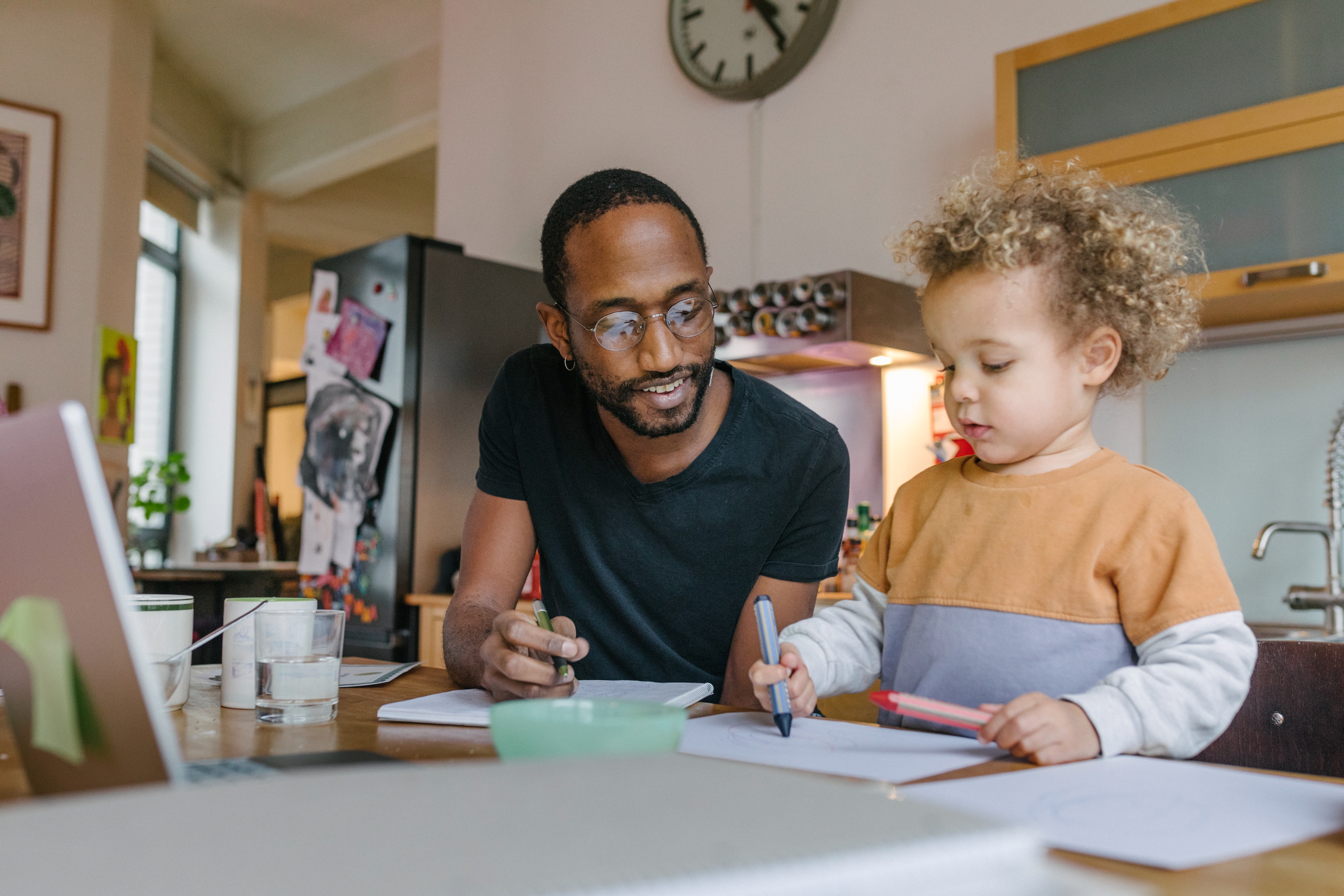 Man and child drawing at a kitchen table, both focused on their papers, surrounded by various art supplies and a laptop