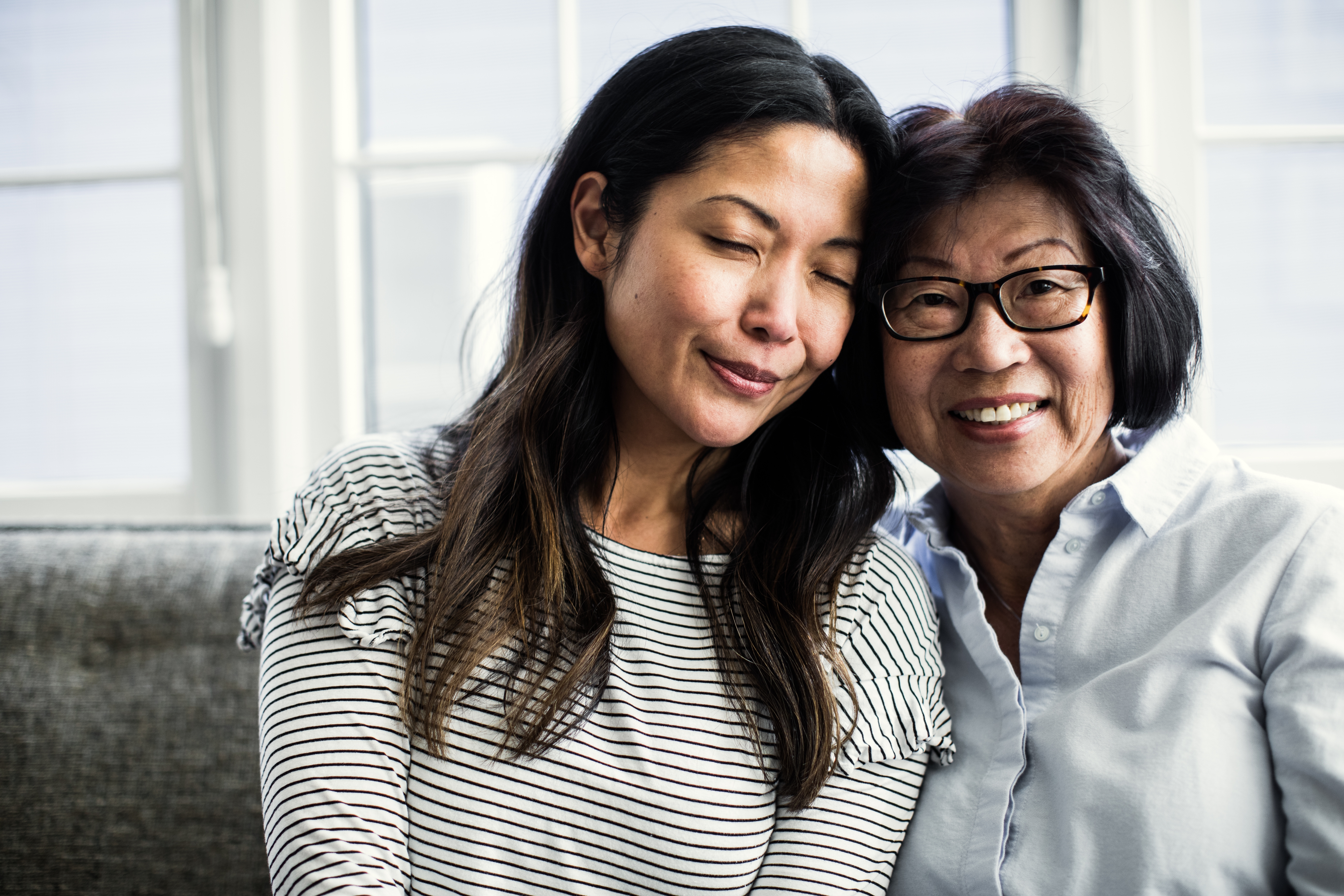 Two women sit closely on a couch, smiling warmly. One wears a striped shirt, the other glasses and a button-down shirt. They appear joyful and relaxed