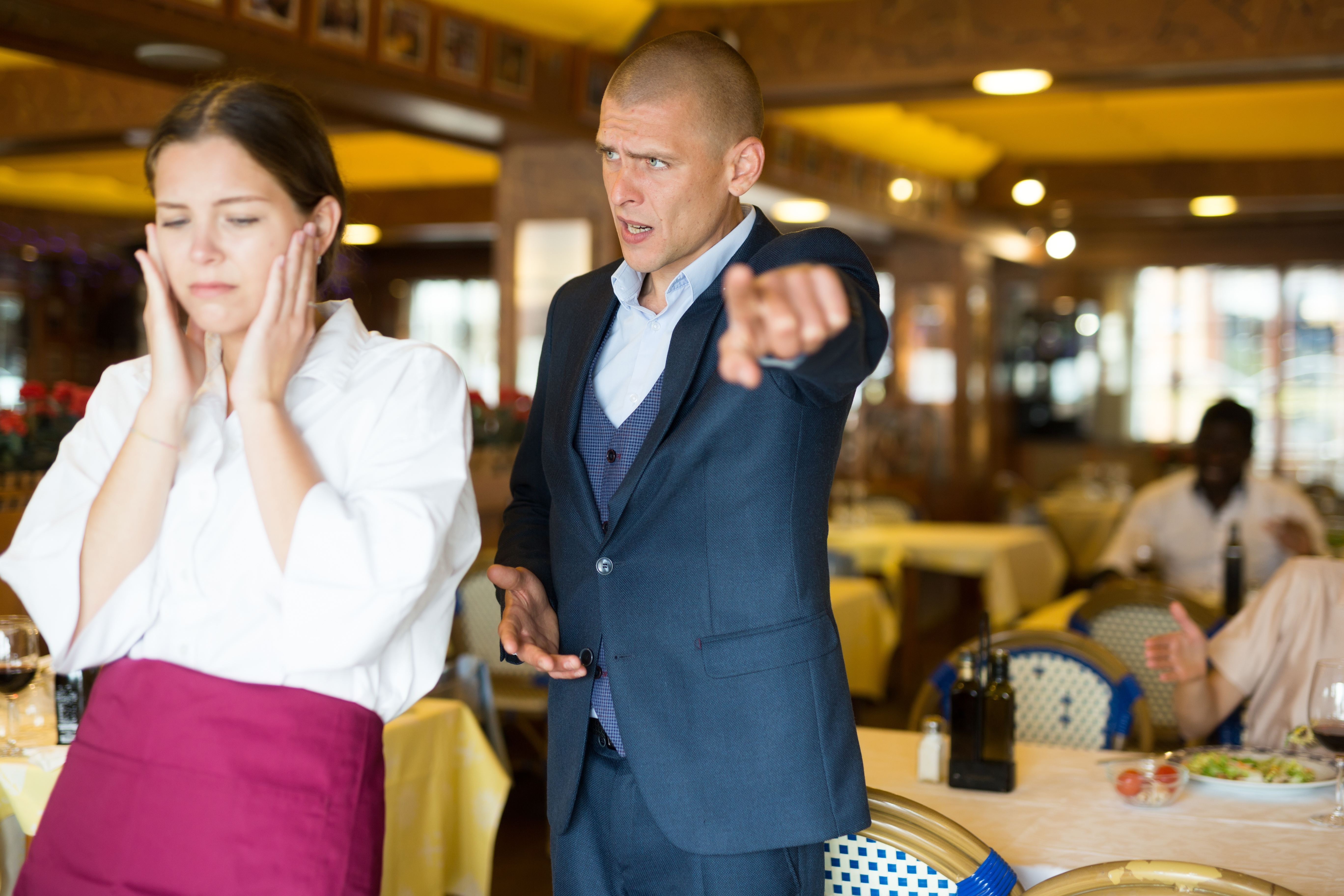 A person in a suit angrily gestures at a server, who looks frustrated, in a restaurant setting