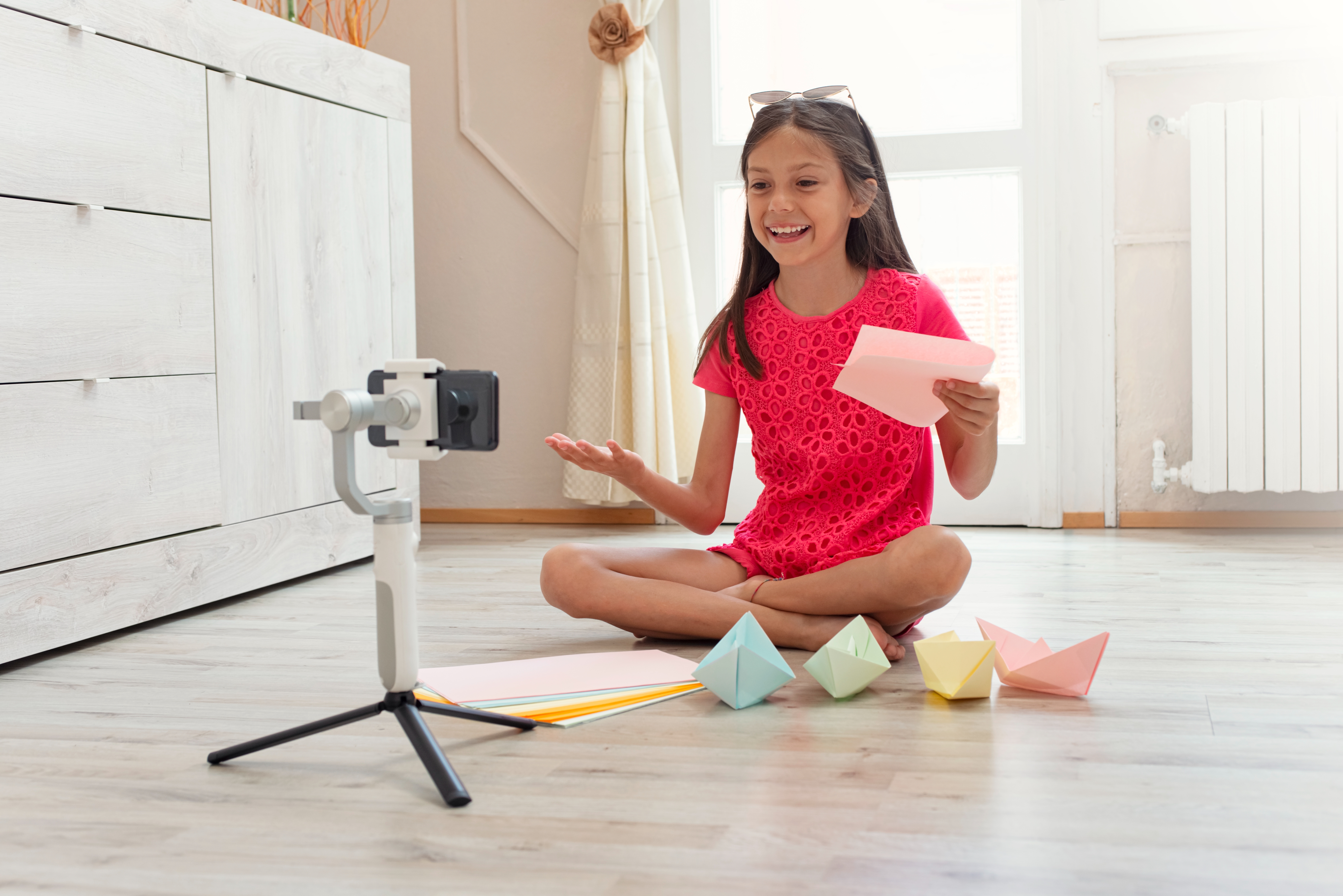 Young girl in a red dress sits cross-legged, making a video with folded paper figures on the floor, smiling and holding notes